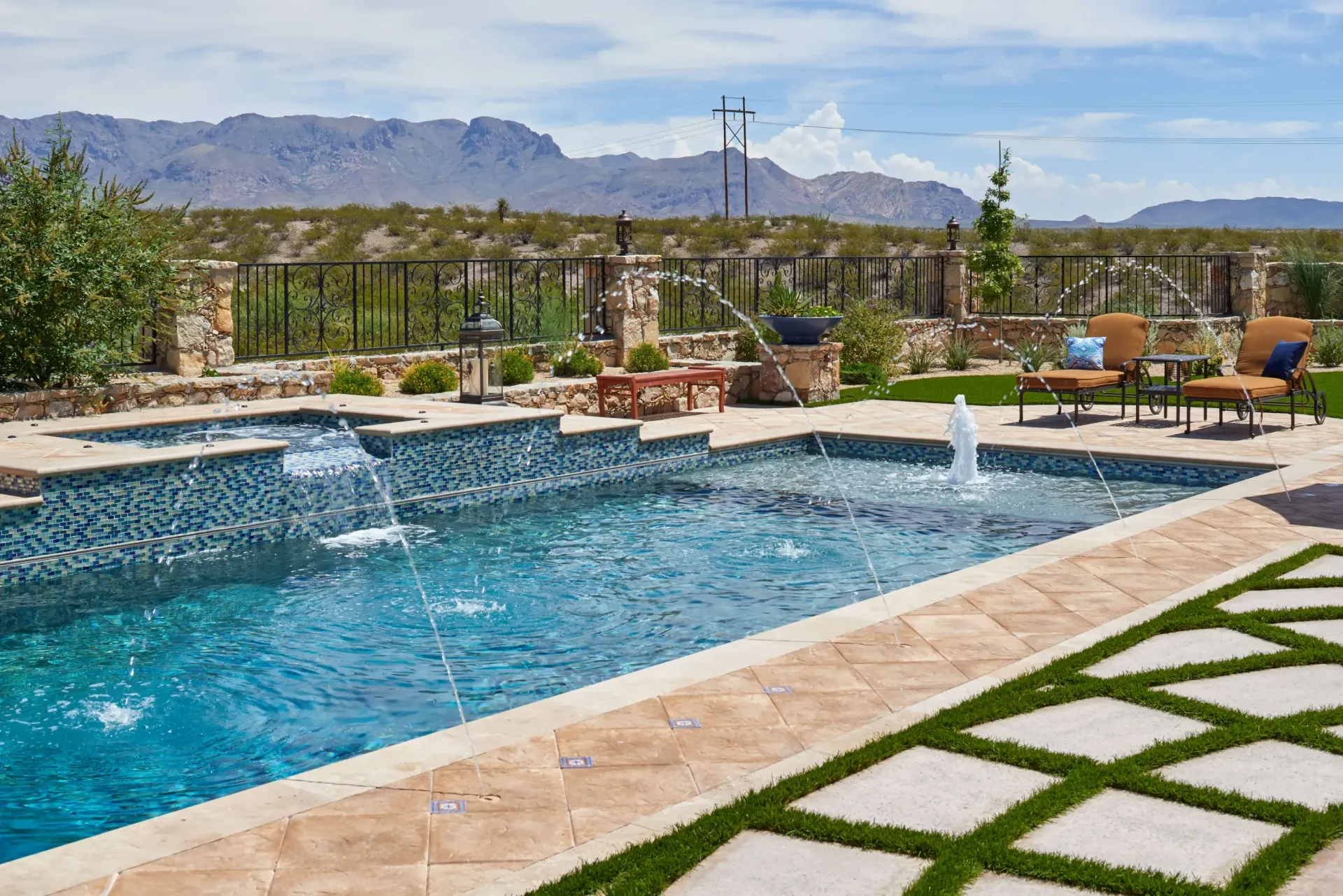 Outdoor swimming pool with water fountains, surrounded by stone and grass with a mountain backdrop.
