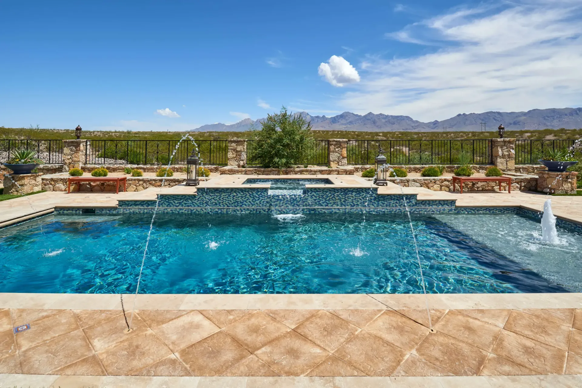 Swimming pool with fountains, blue water, tan stone patio, and mountain view under a blue sky.