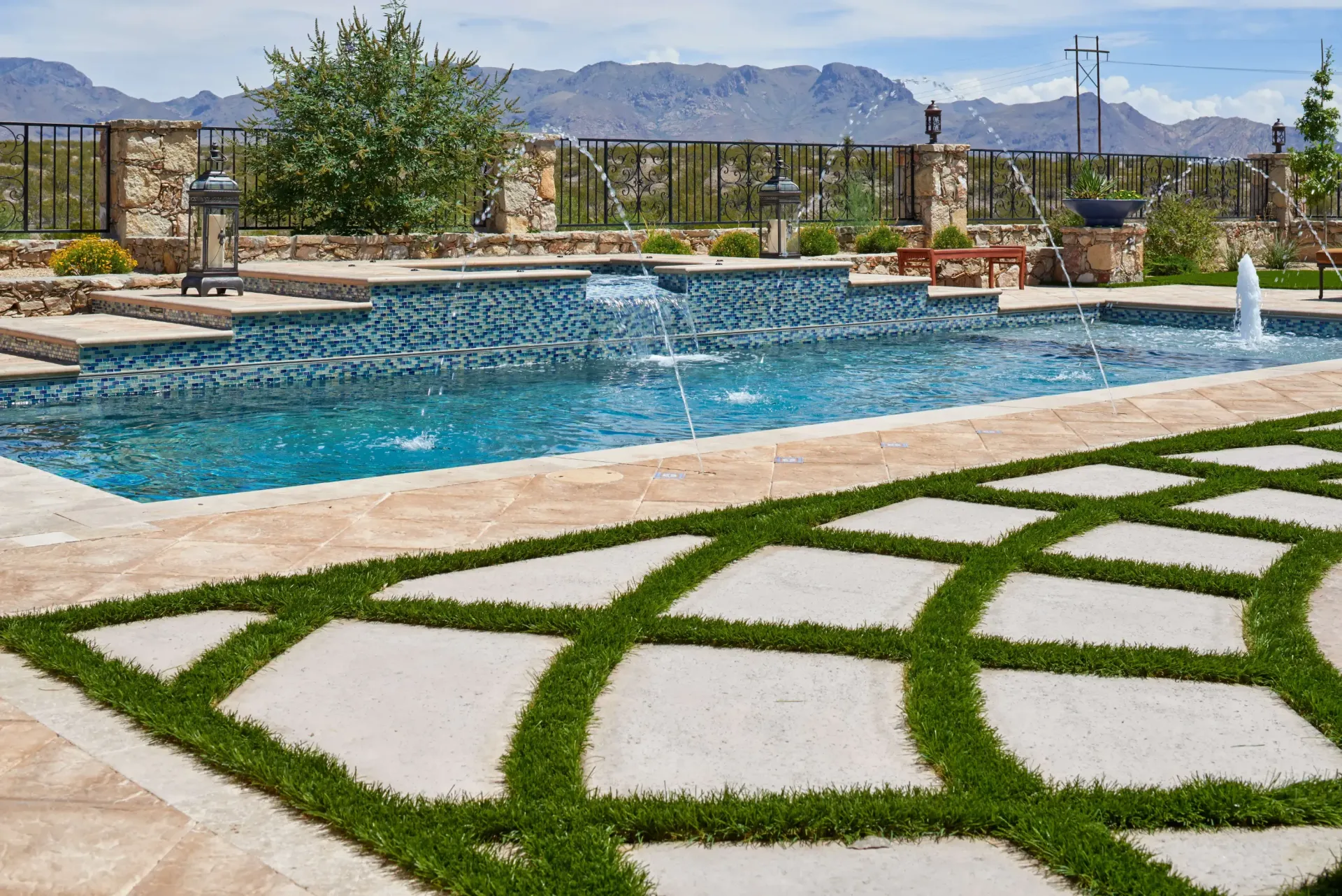 Swimming pool with fountains and blue tile, surrounded by stone patio and grassy paths, mountains in background.