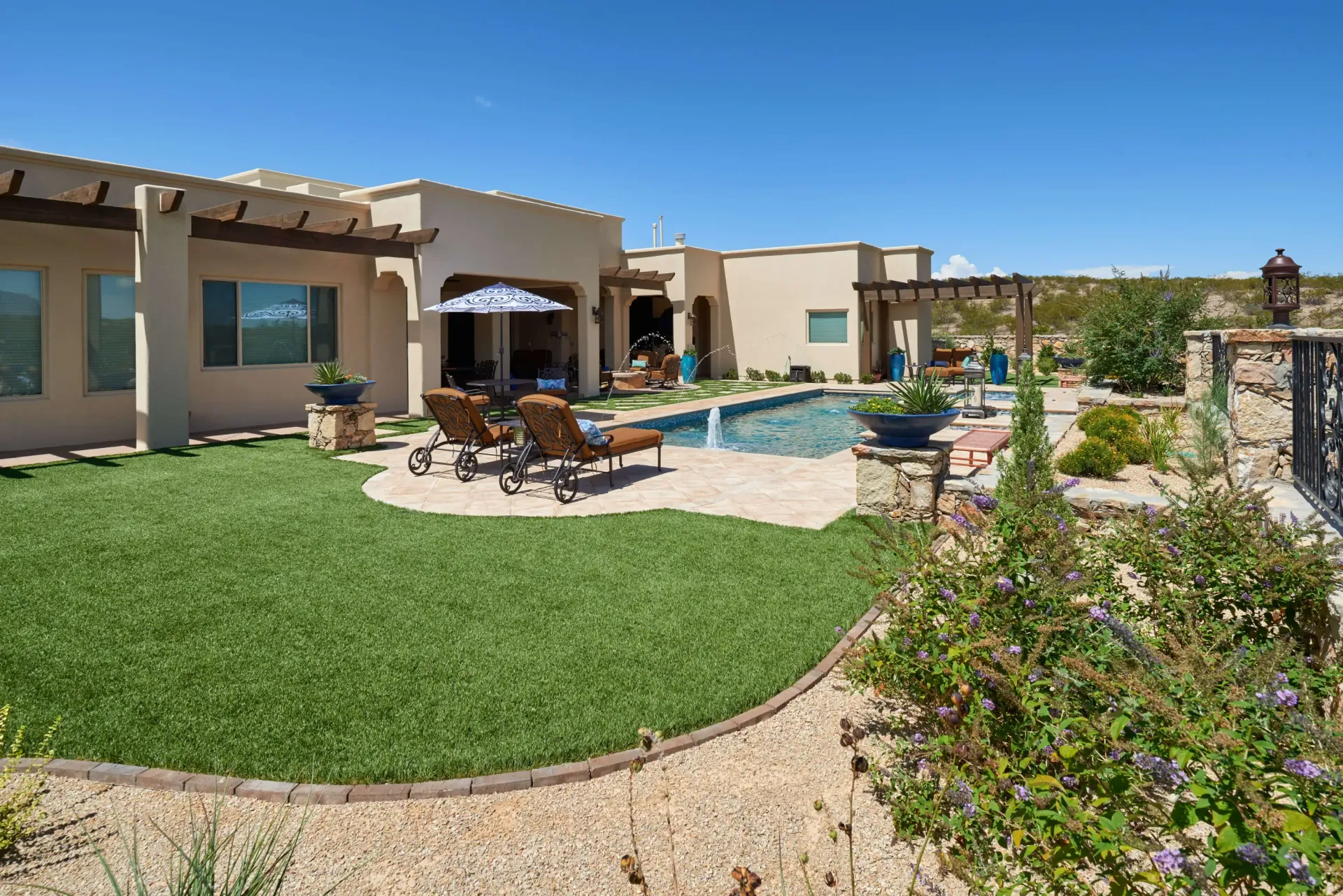 Backyard with pool, patio, and lawn. Tan house, blue umbrella, lounge chairs, and desert landscape.