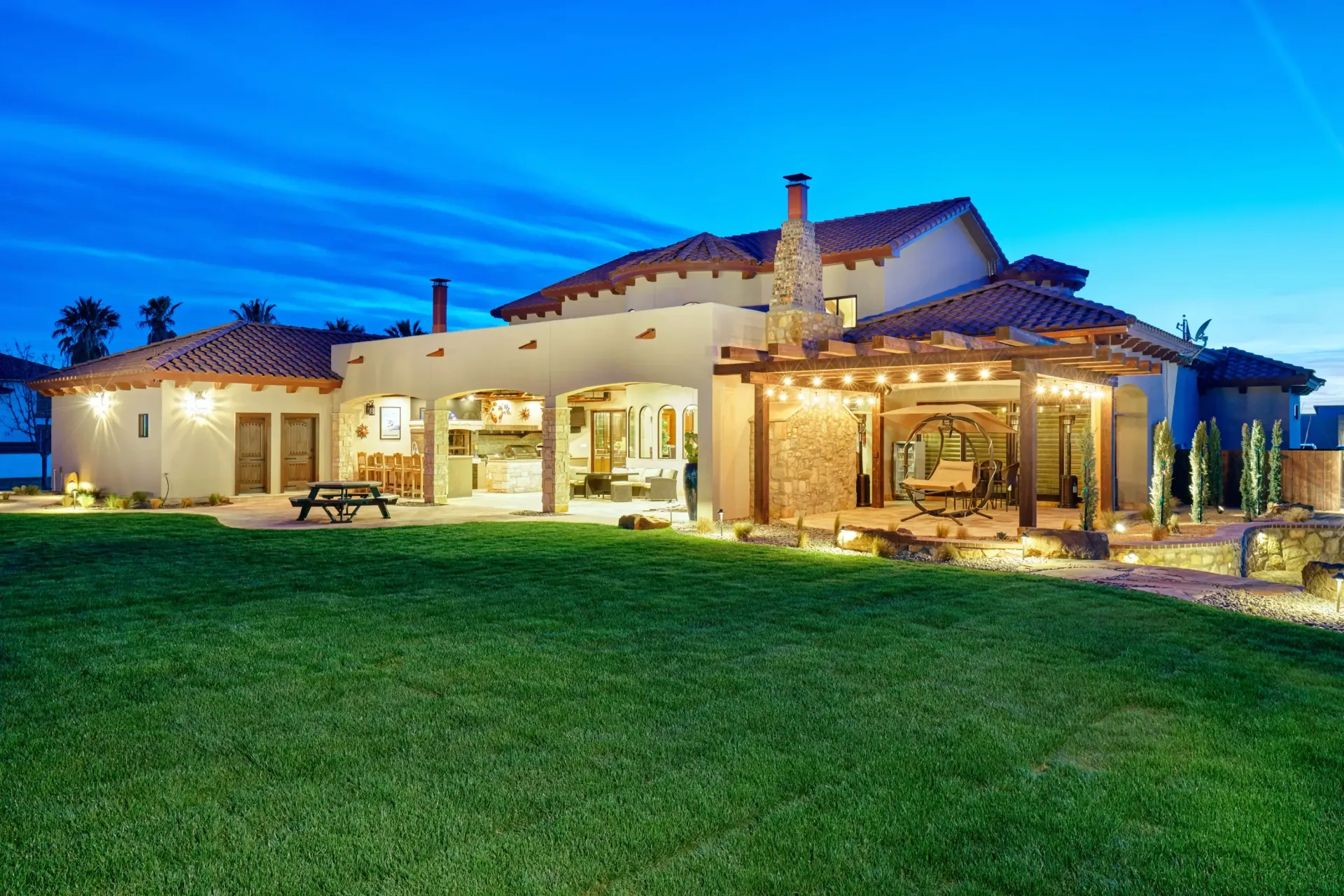 Backyard view of a stucco house with tiled roof and covered patio, illuminated at dusk, with lush green lawn.