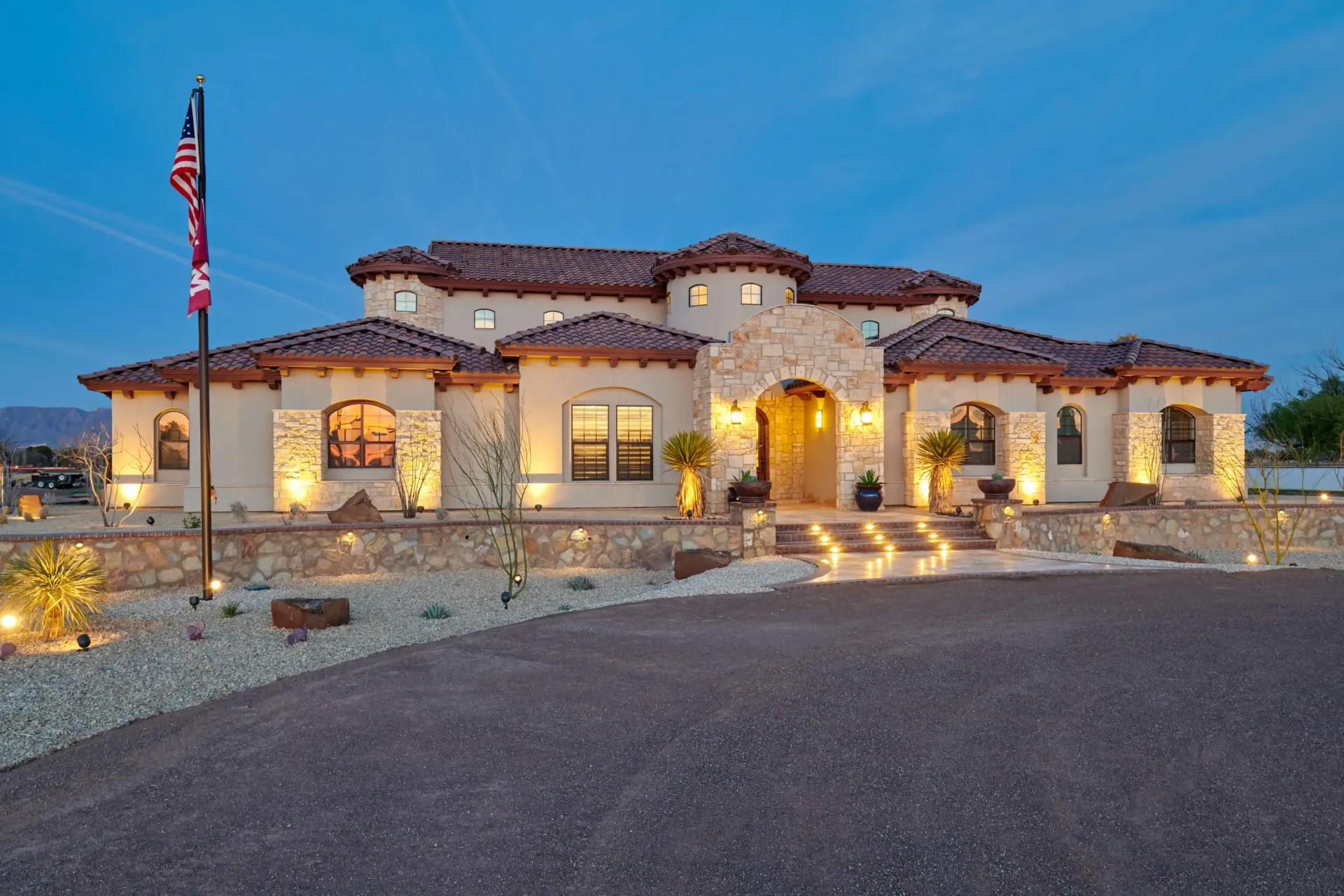 Large, stucco-walled home with red-tile roof at dusk, featuring a circular driveway, flag, and illuminated entry.