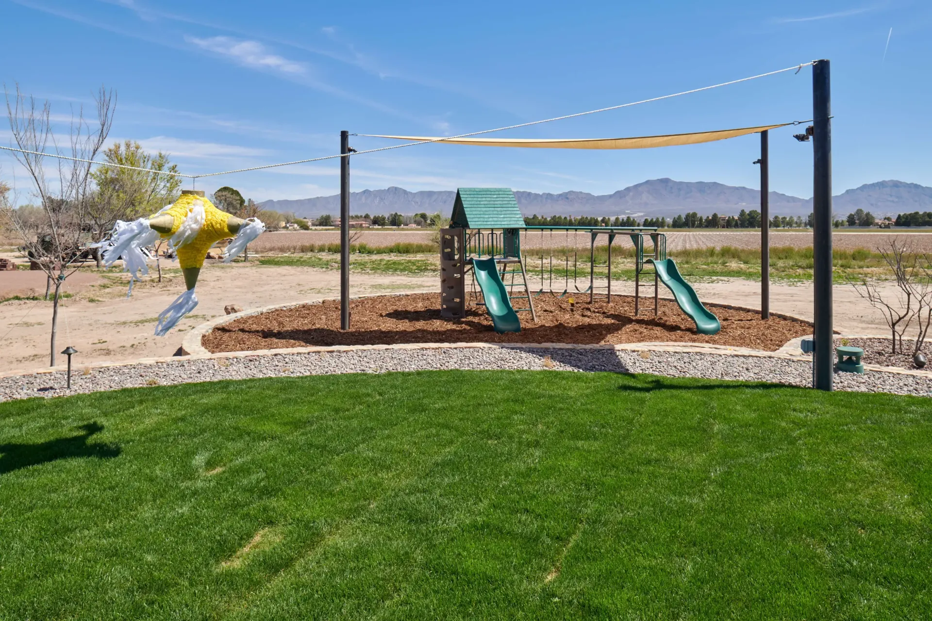 Playground with slides and swing set, under a sun shade, with a green lawn. Mountains in background.