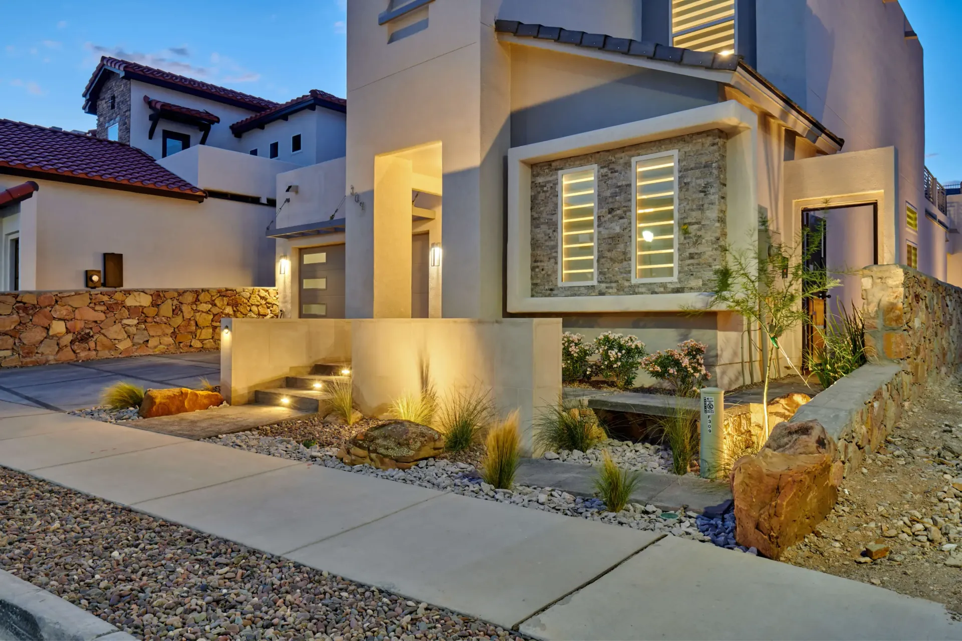 Modern two-story house with illuminated landscaping, stone retaining walls, and a driveway at dusk.