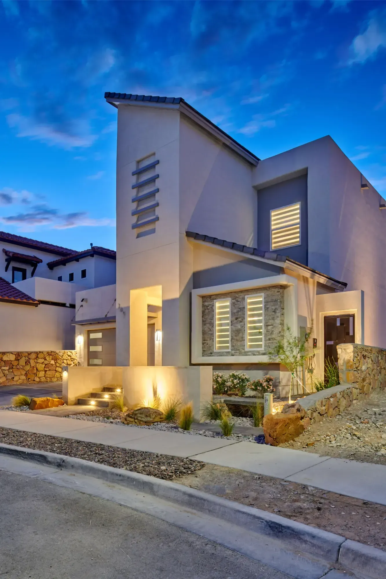 Modern two-story house with stucco exterior, at dusk. Features include angular design, stone accents, and outdoor lighting.