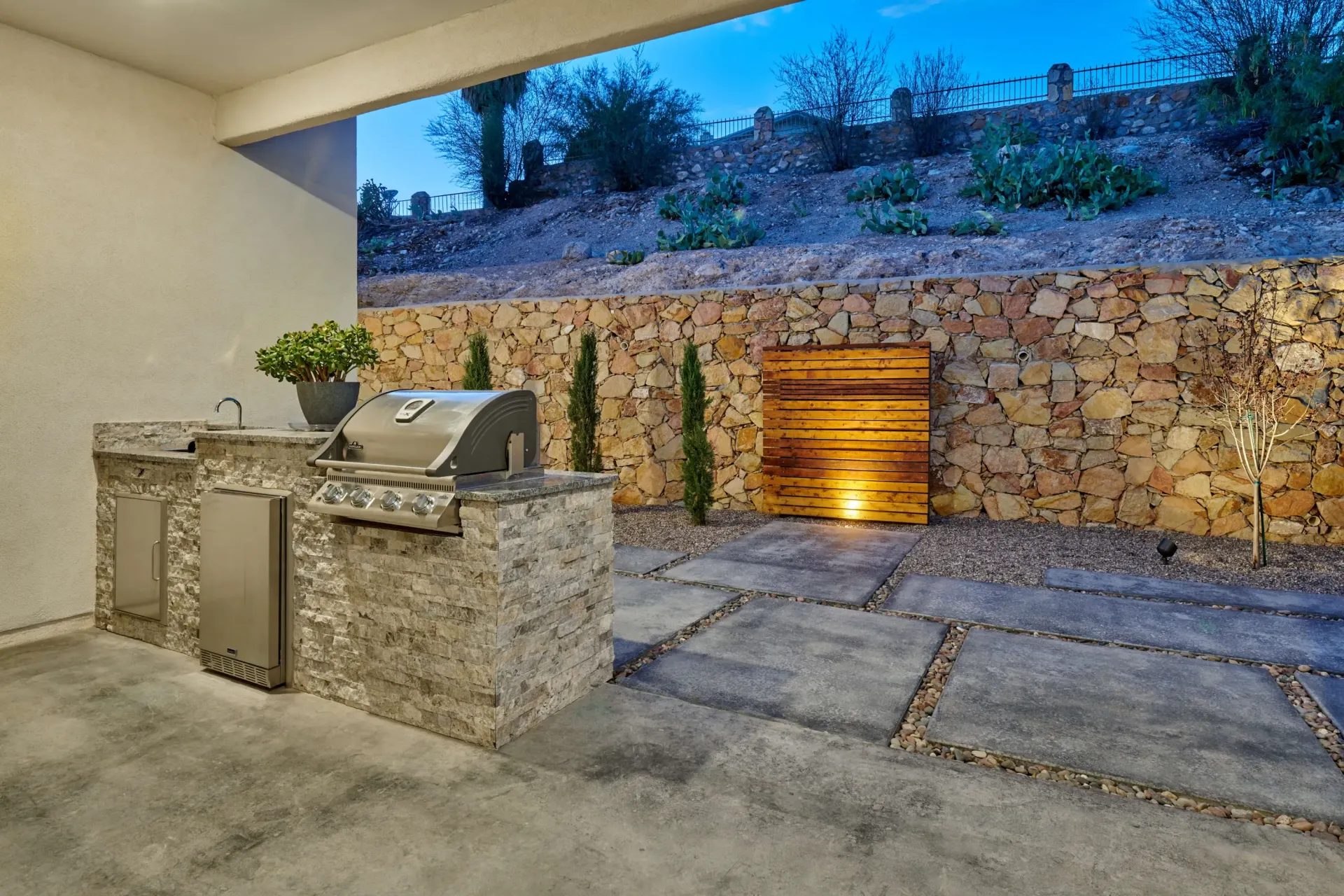 Outdoor kitchen with built-in grill, stone walls, and a wood accent, under an awning, at dusk.