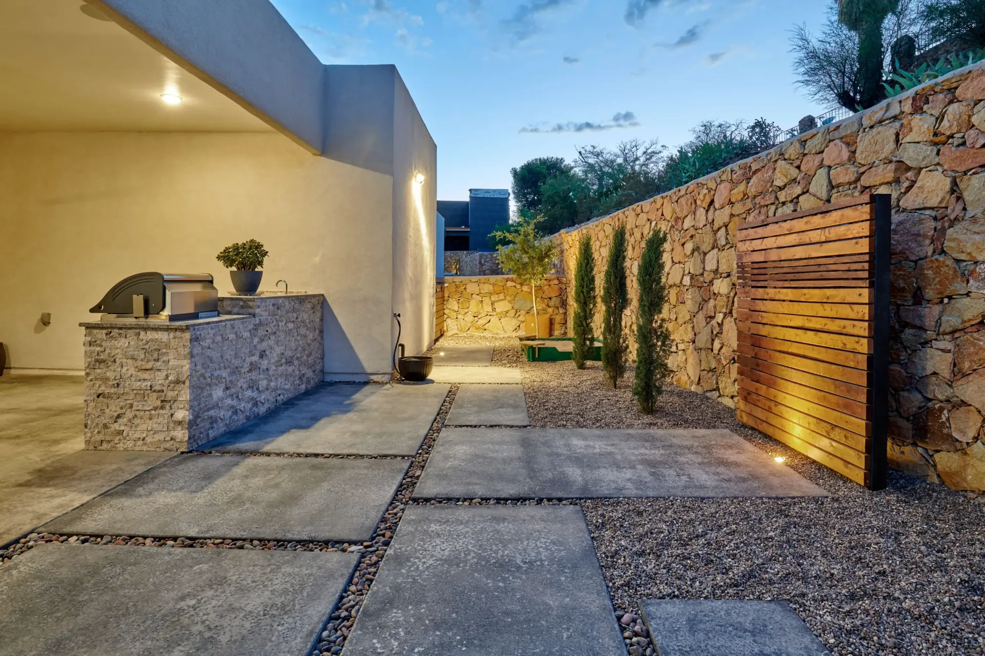 Patio with a stone wall and pathway, featuring a built-in grill and wooden gate at dusk.