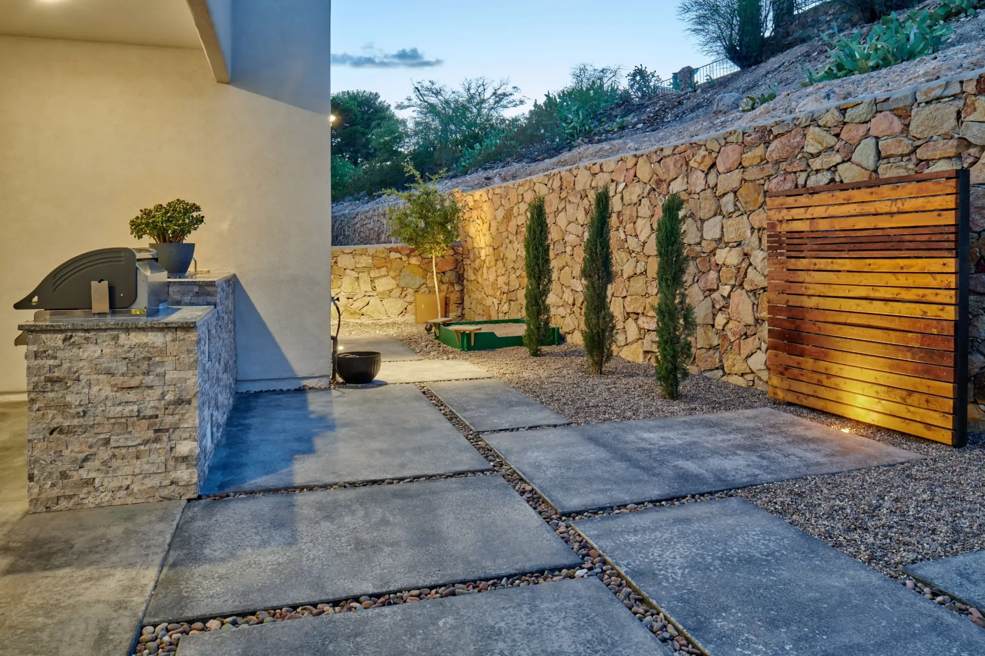 Patio with stone wall, grill, square concrete pavers, gravel, and wood paneling, lit by warm light.