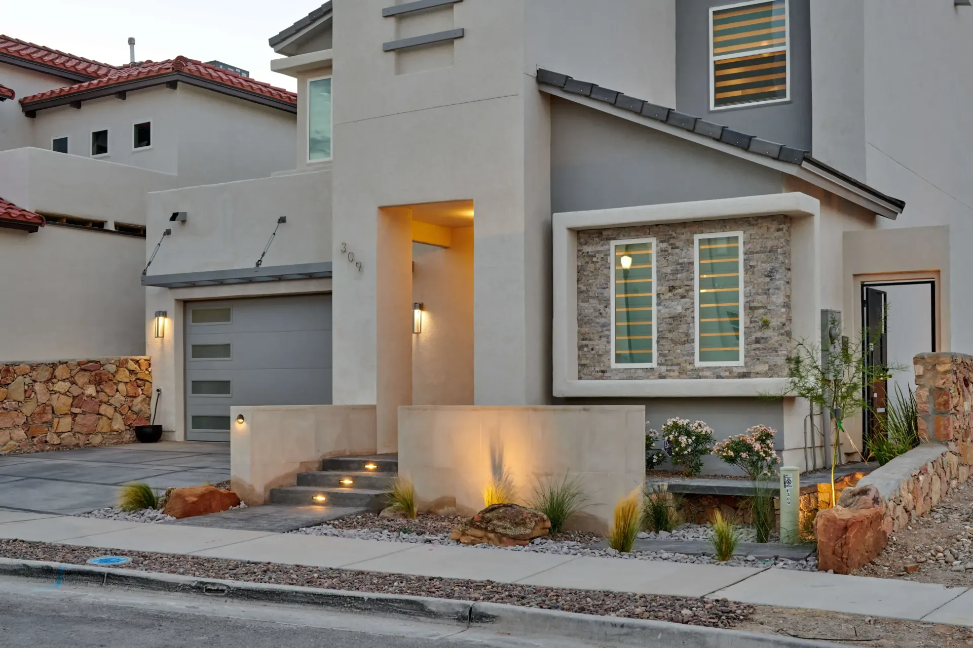 Modern two-story house with gray and tan stucco exterior, stone accents, and illuminated entrance stairs.