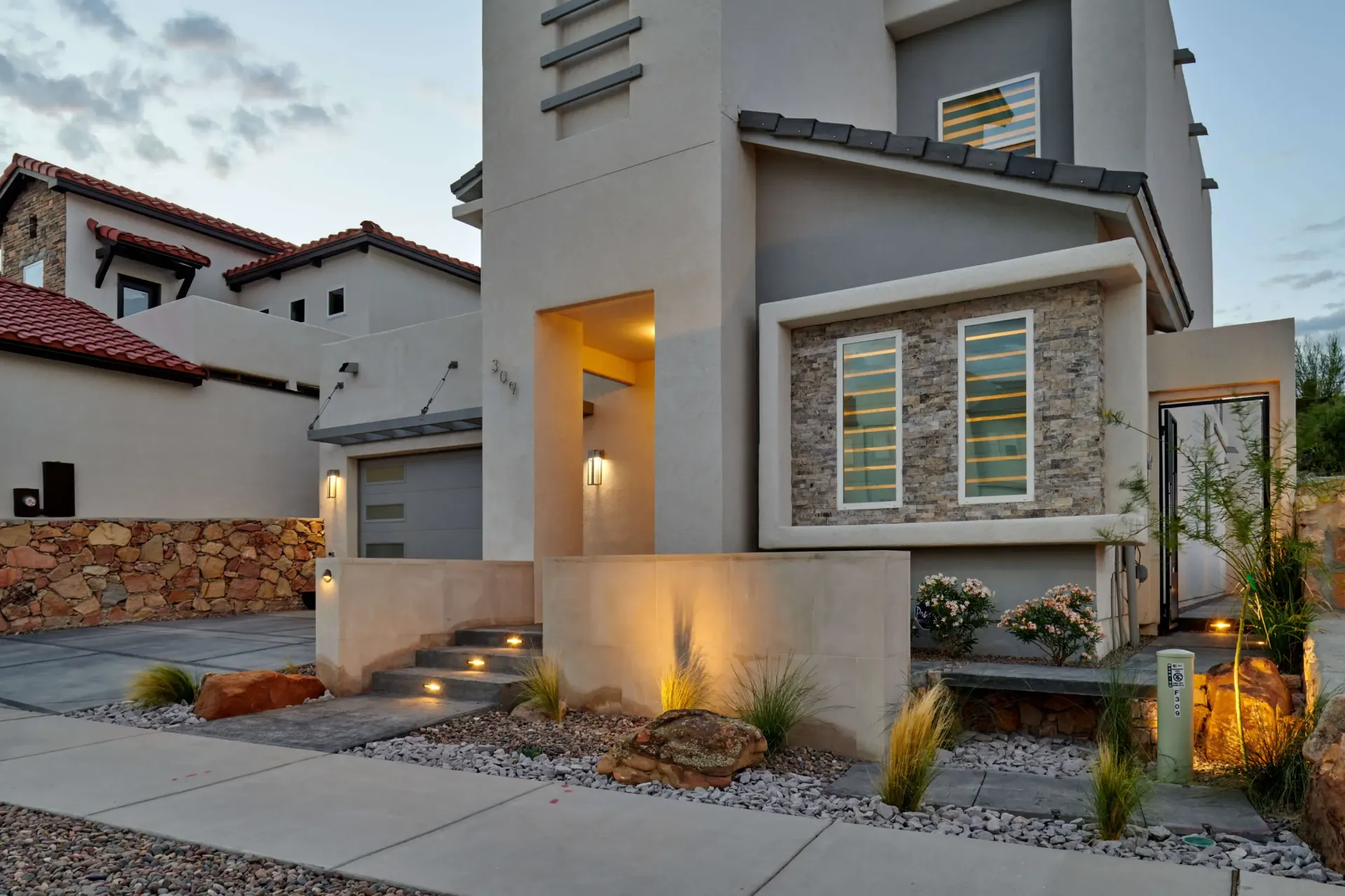 Modern two-story house with gray and tan stucco exterior, stone accents, and low-lit landscaping.