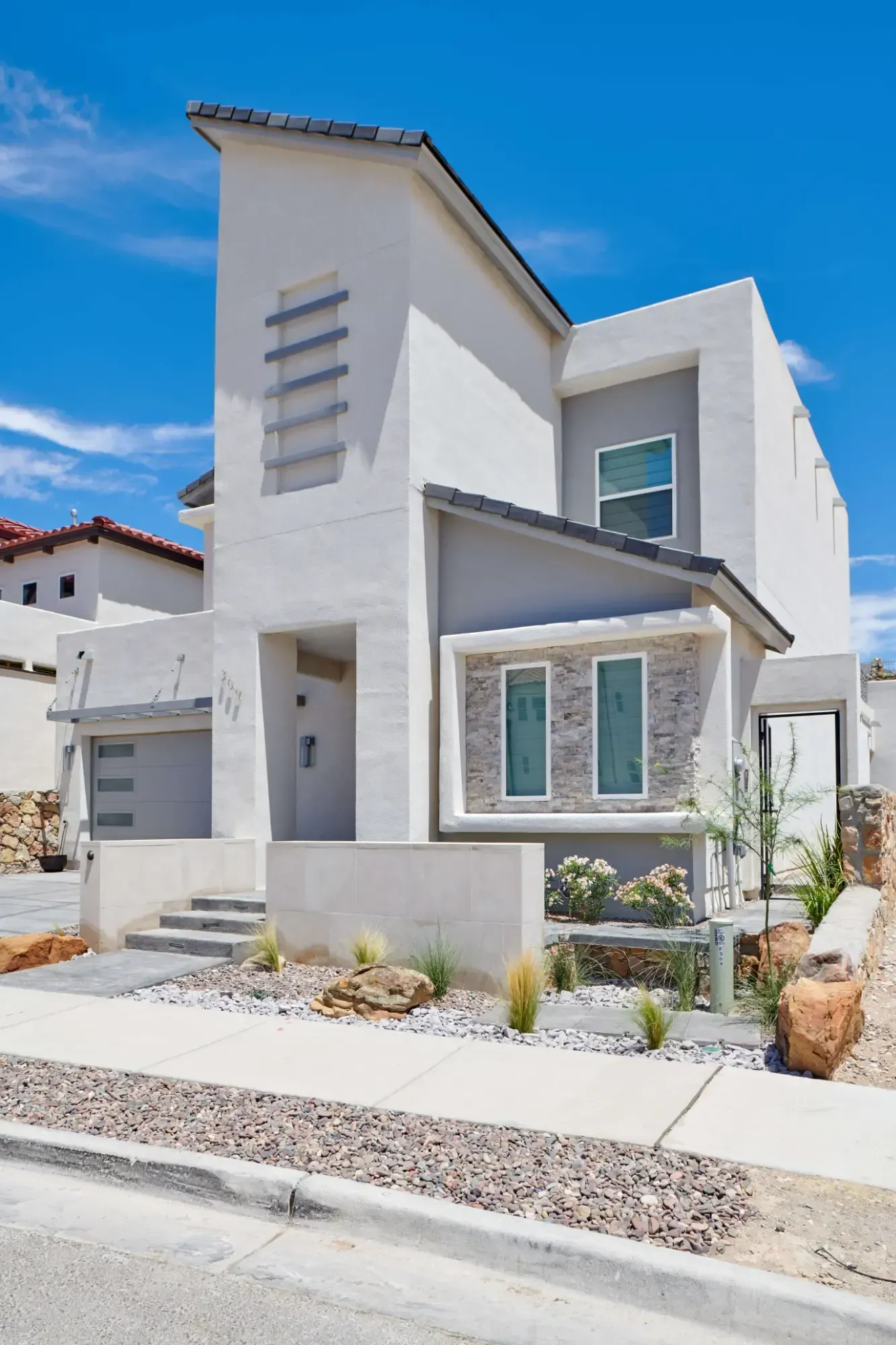 Modern two-story house with gray and white exterior, blue sky background. Landscaping with small plants and rocks in front.