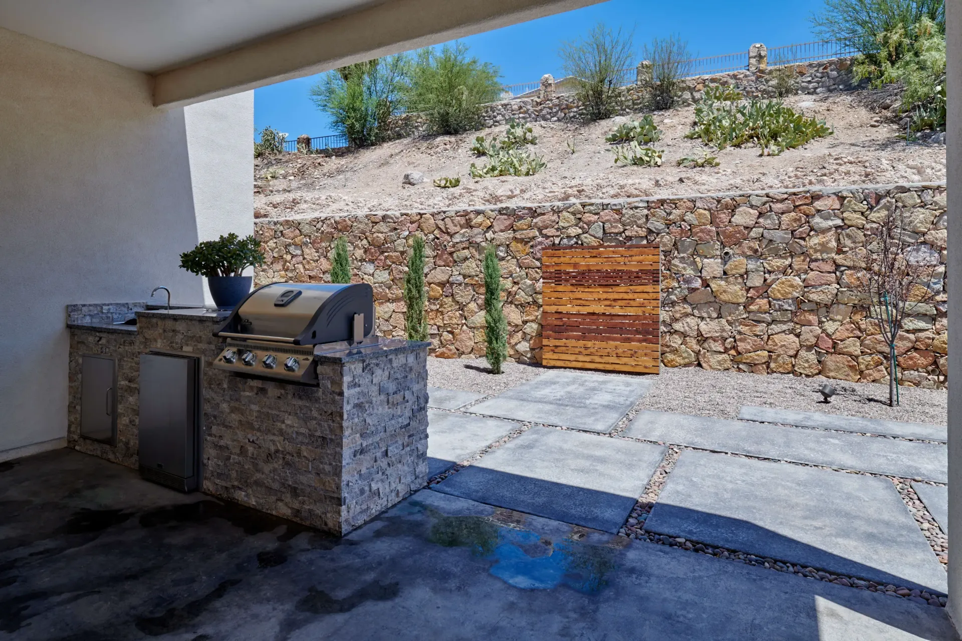 Outdoor kitchen with grill, sink, and stone countertop; patio with a stone wall and landscaping.