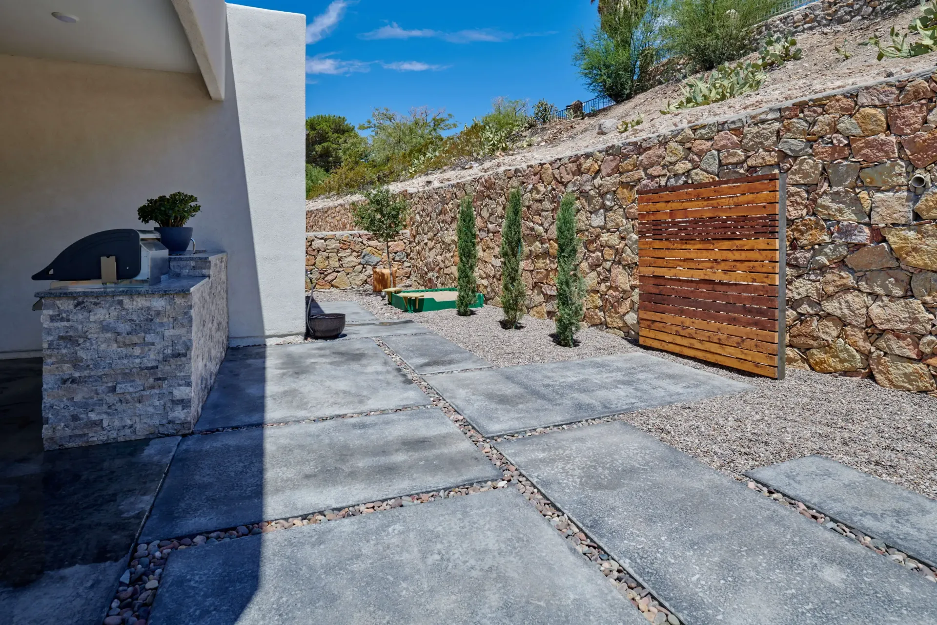 Backyard patio with stone grill, concrete pavers, gravel, and tiered retaining wall.