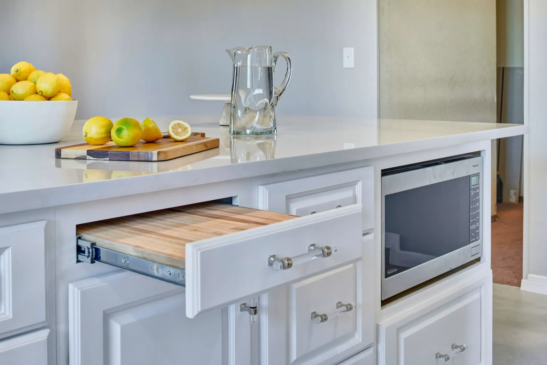 Kitchen island with pull-out cutting board, lemons, pitcher, and built-in microwave. White cabinets and countertop.