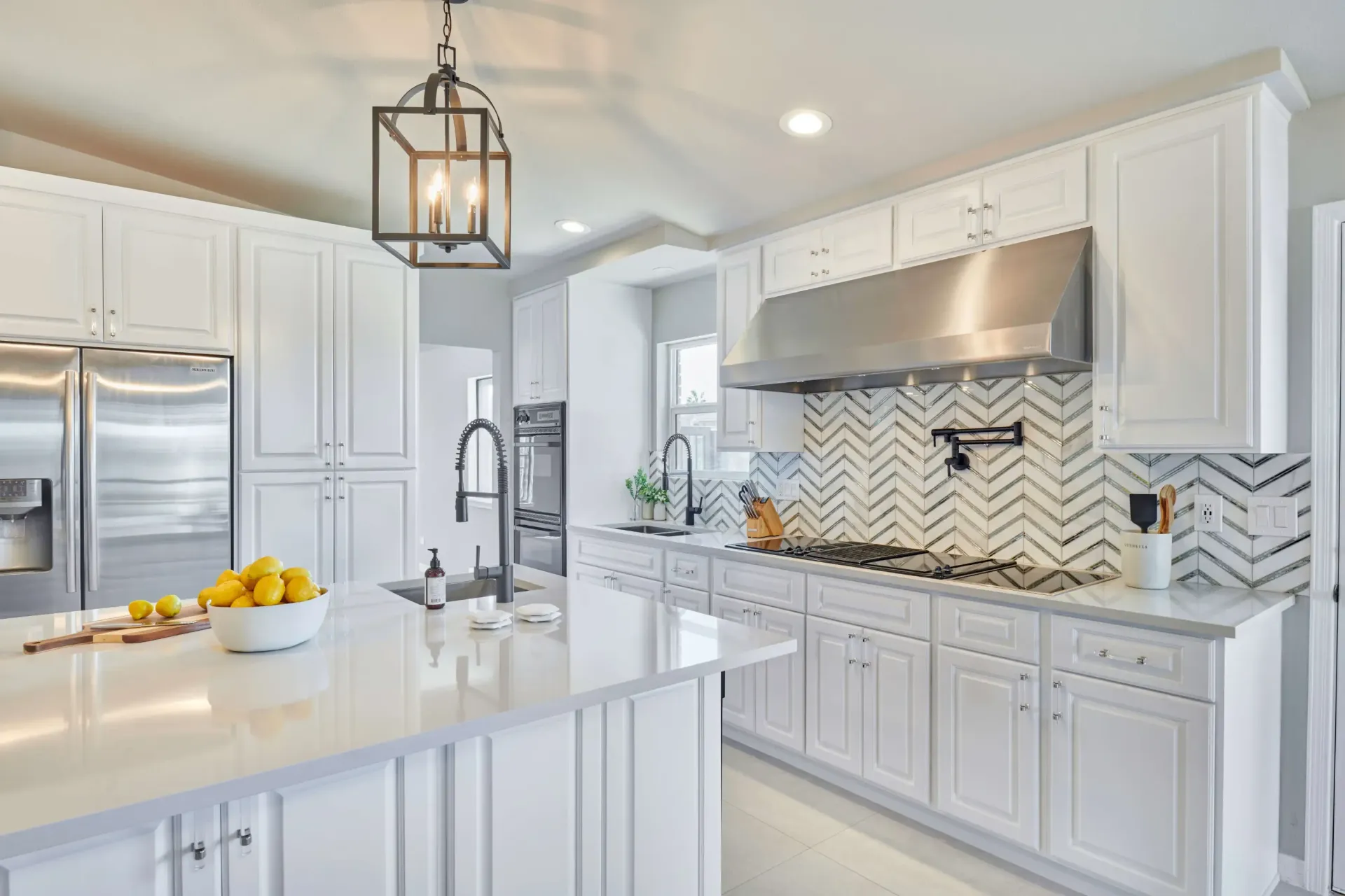 White kitchen with island, stainless steel appliances, herringbone backsplash, and pendant light.
