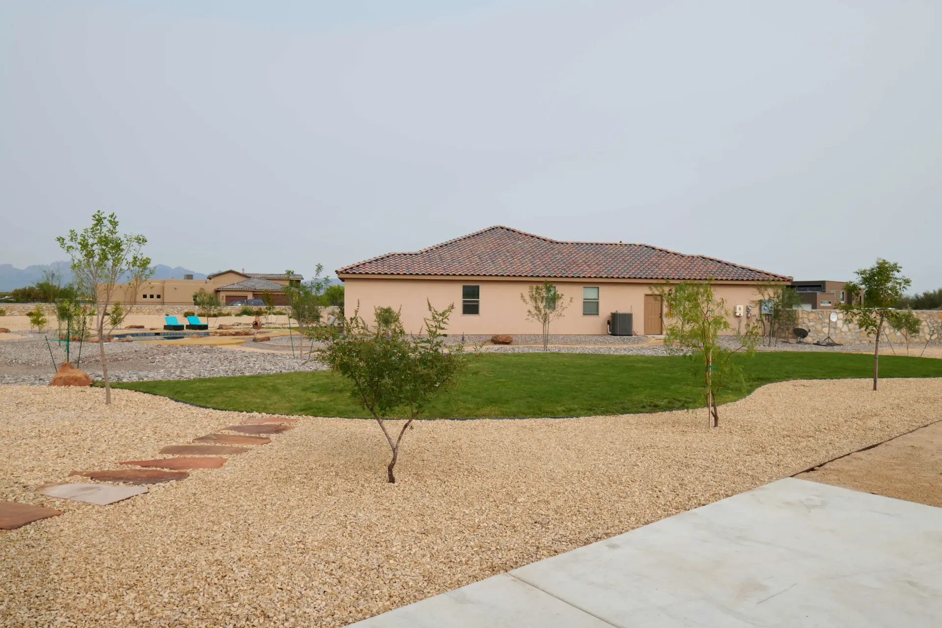 Tan house with tile roof, surrounded by tan gravel and sparse green grass, under an overcast sky.