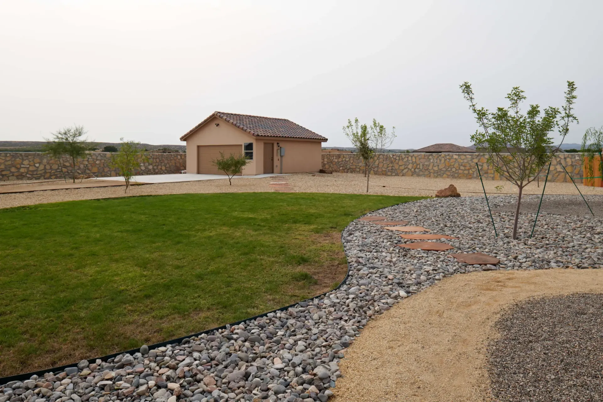 A tan stucco garage with a red-tiled roof sits on a concrete pad surrounded by gravel, grass, and young trees.