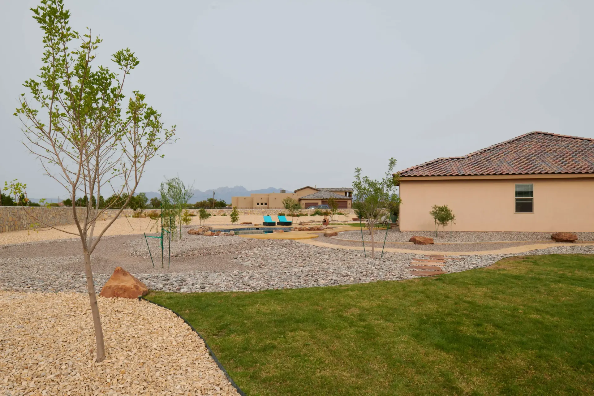 A home's xeriscaped backyard with a lawn, gravel, trees, and a tan house under an overcast sky.