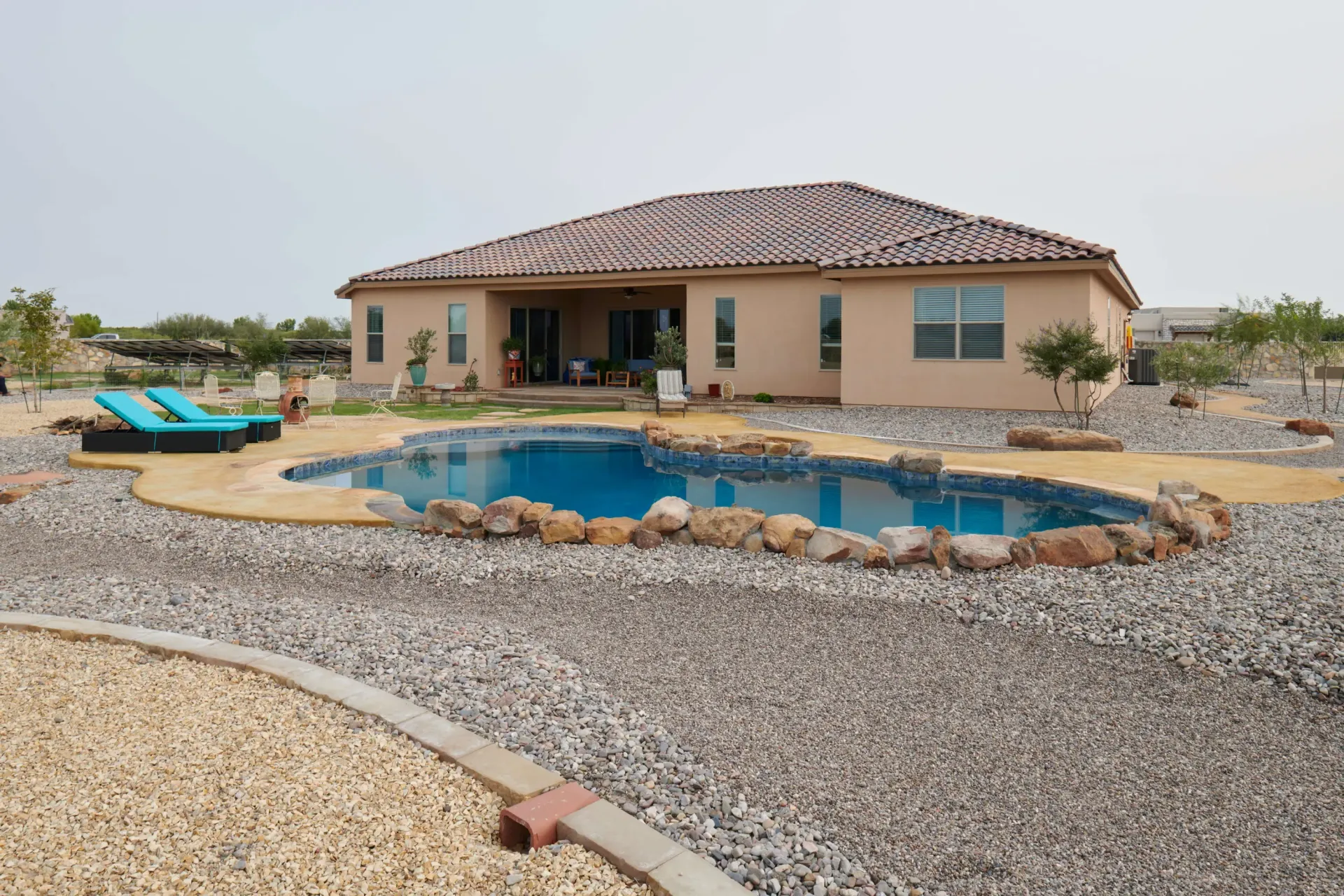 A house with a pool in a yard covered in gravel. Blue pool with lounge chairs, tan house, and brown roof.