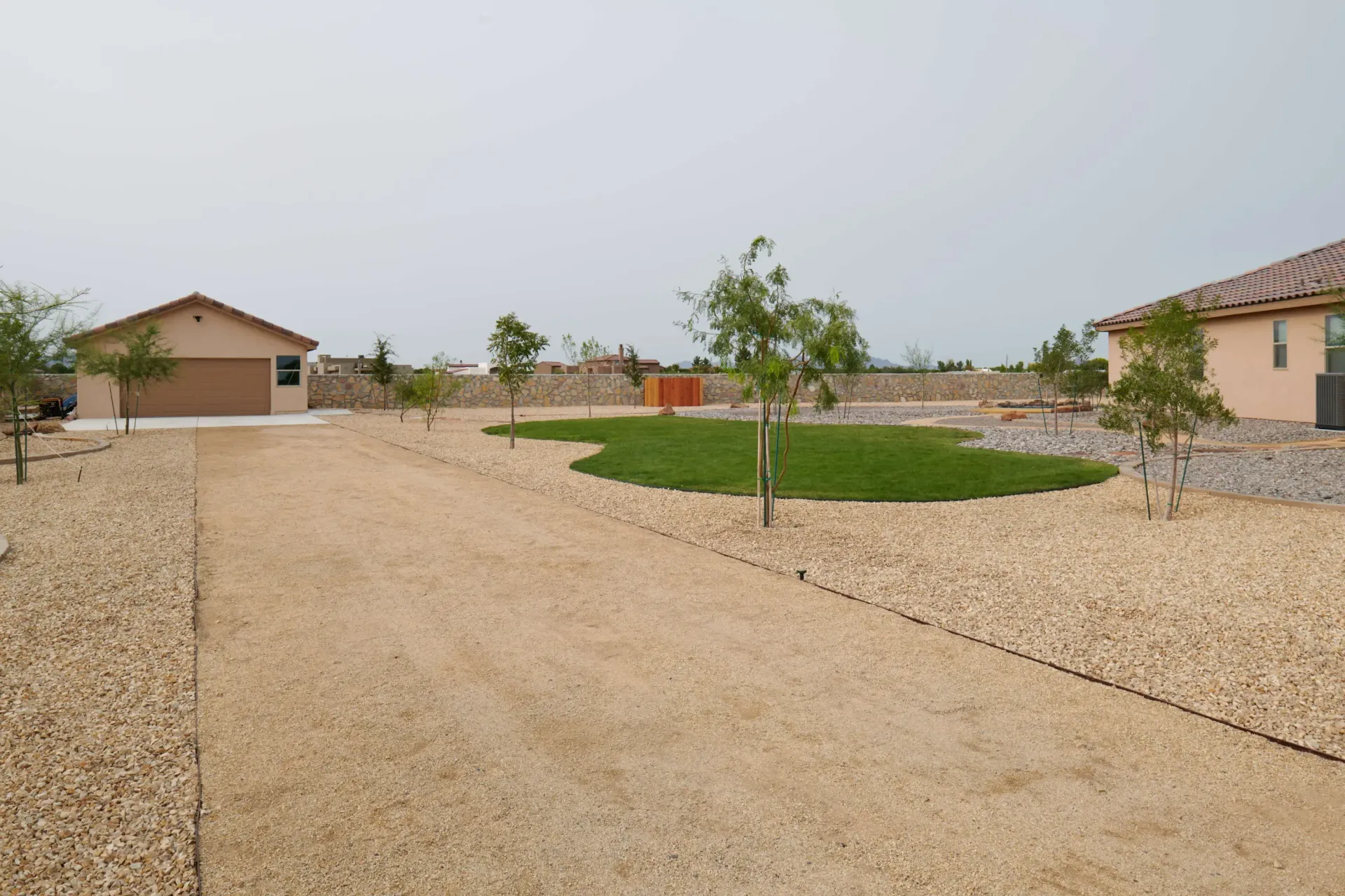 Gravel driveway leading to a garage and house, small trees, and a grassy area under an overcast sky.