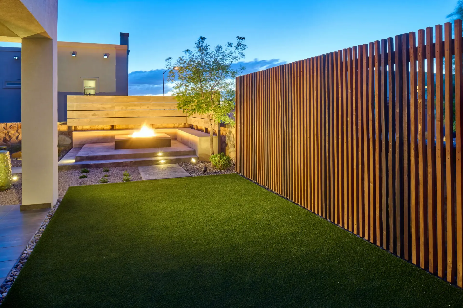 Backyard with artificial grass, a fire pit, wooden slatted fence, and a modern home.