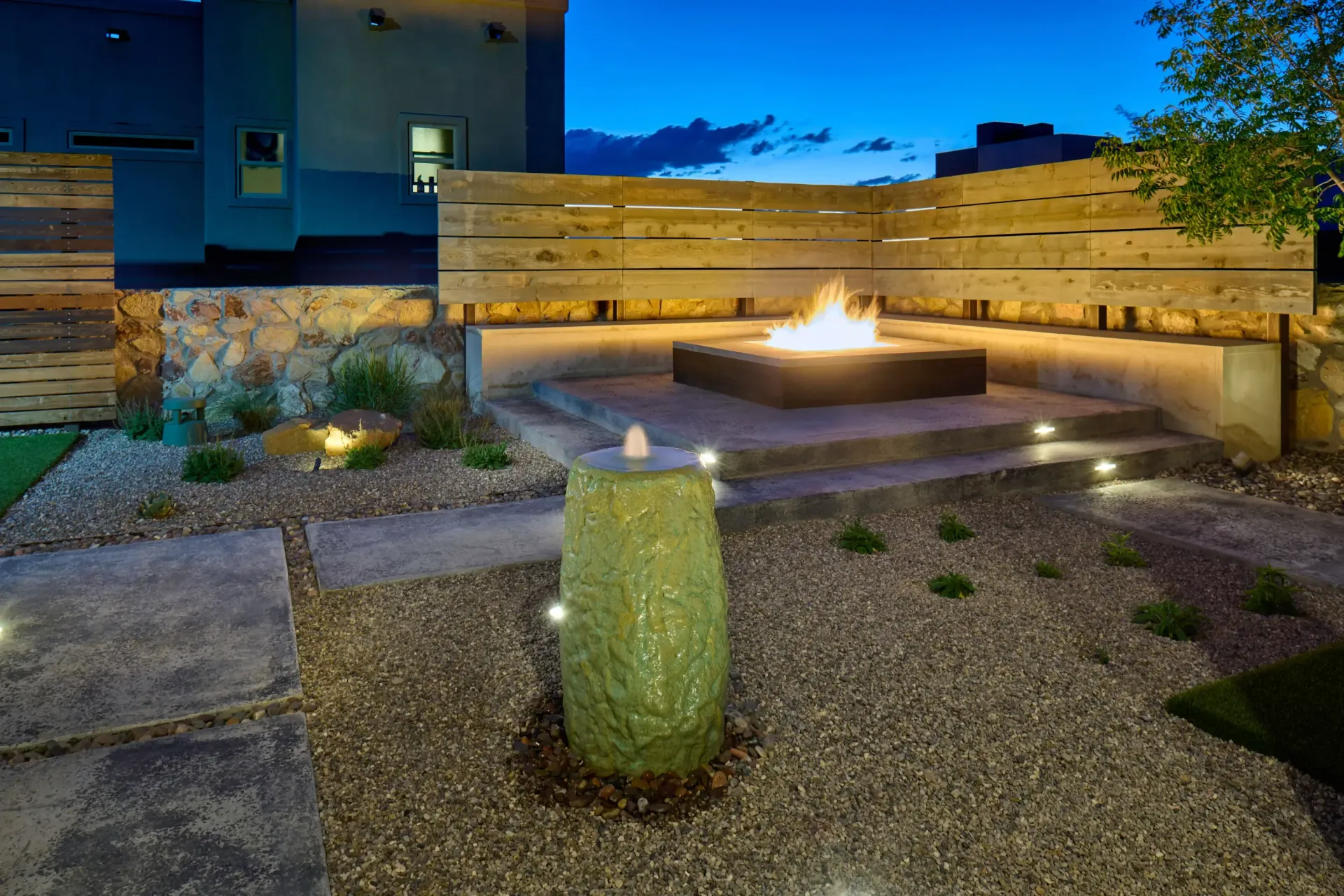 Nighttime outdoor patio with fire pit, illuminated fountain, and wood fence.