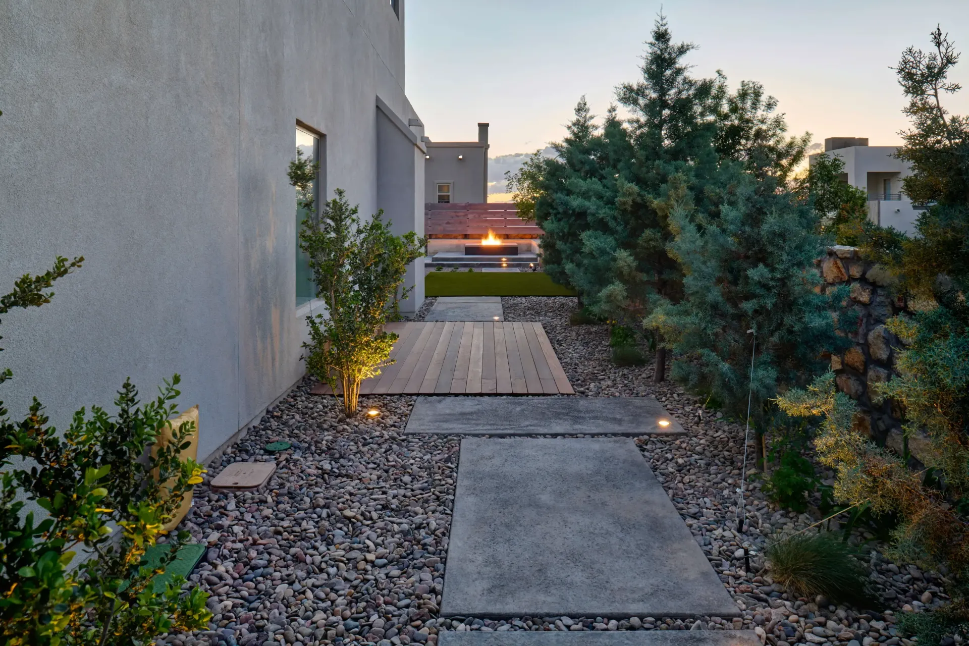 Stone pathway lined with gravel and greenery leads to a patio with a fire pit at dusk.