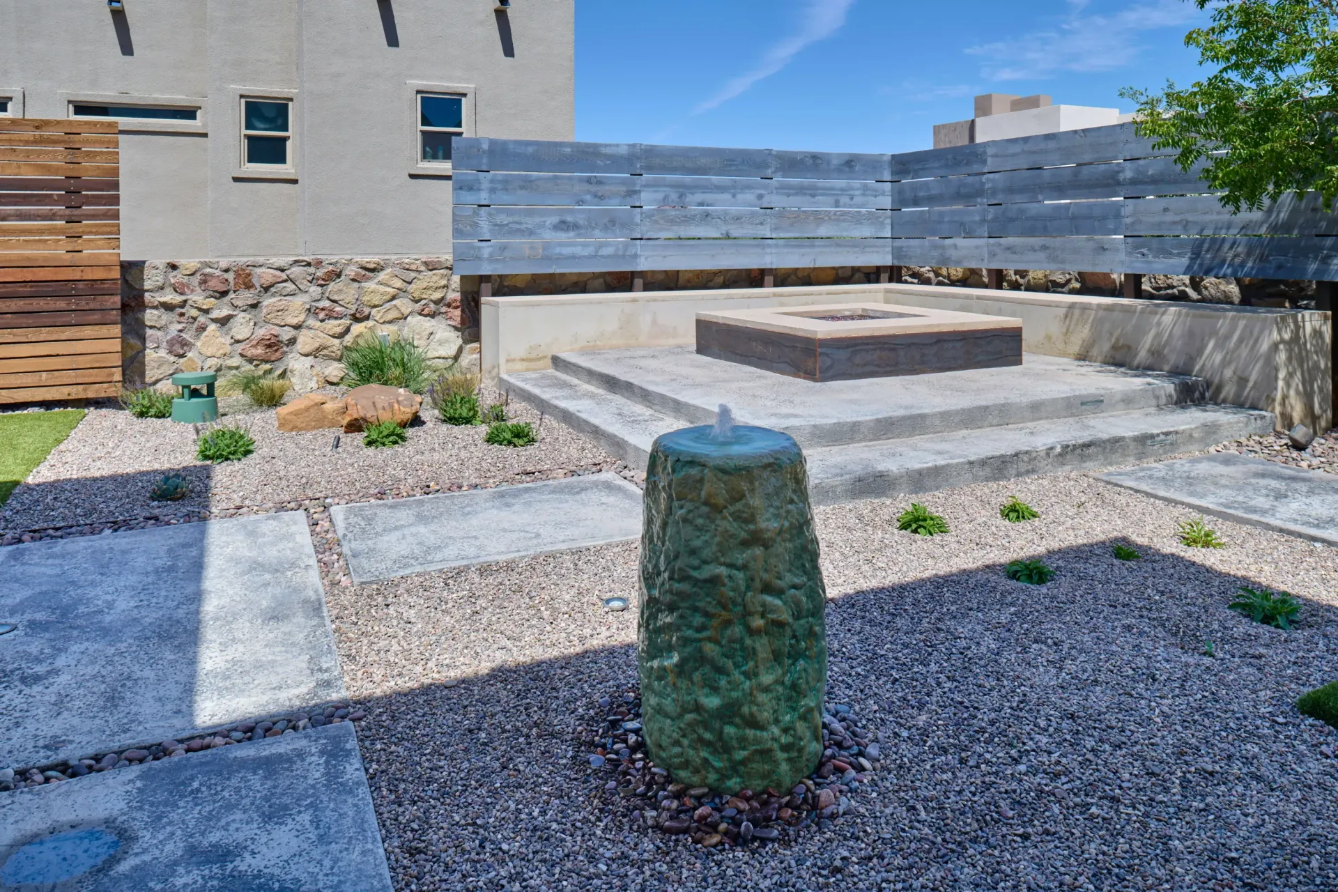 Stone water fountain in a gravel yard with a concrete patio, fire pit, and wooden fence.