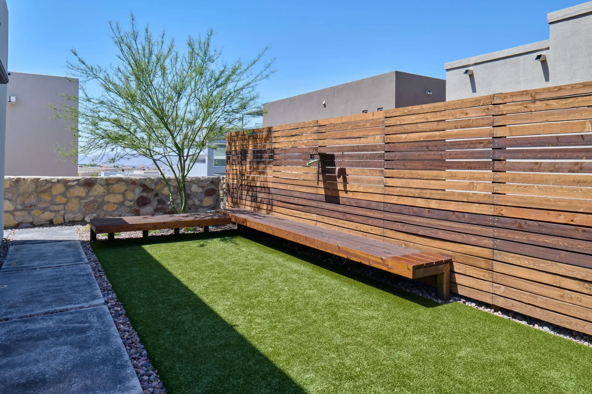 Rooftop patio with artificial turf, wooden benches, a tree, and a wooden fence on a sunny day.
