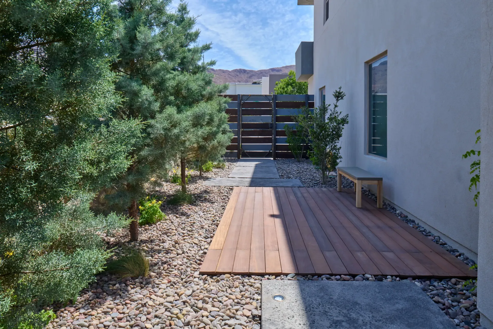 Pathway with wood deck, bench, trees, and fence next to a white building.