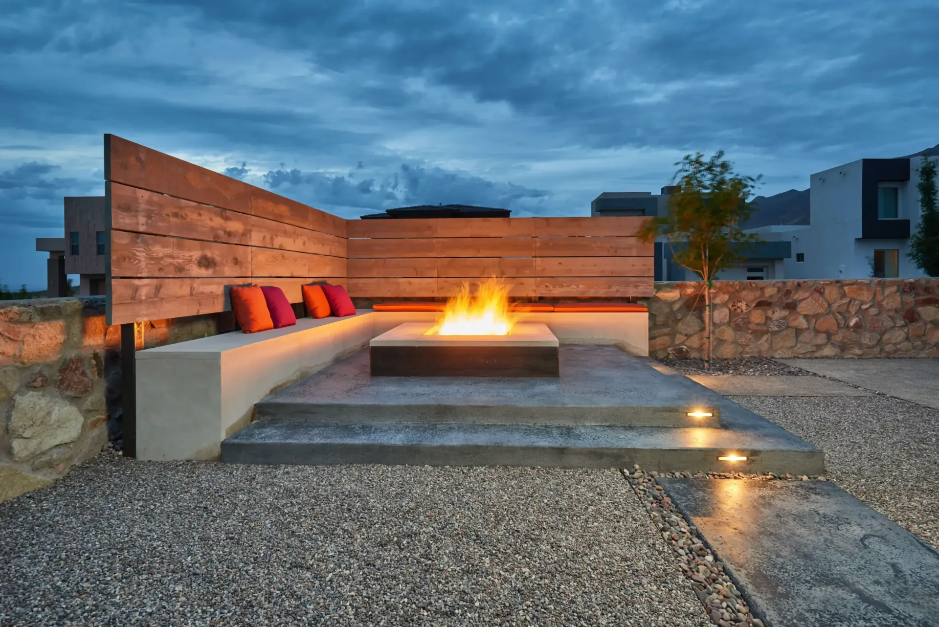 Outdoor patio with fire pit, concrete seating, and warm lighting under a dusk sky.
