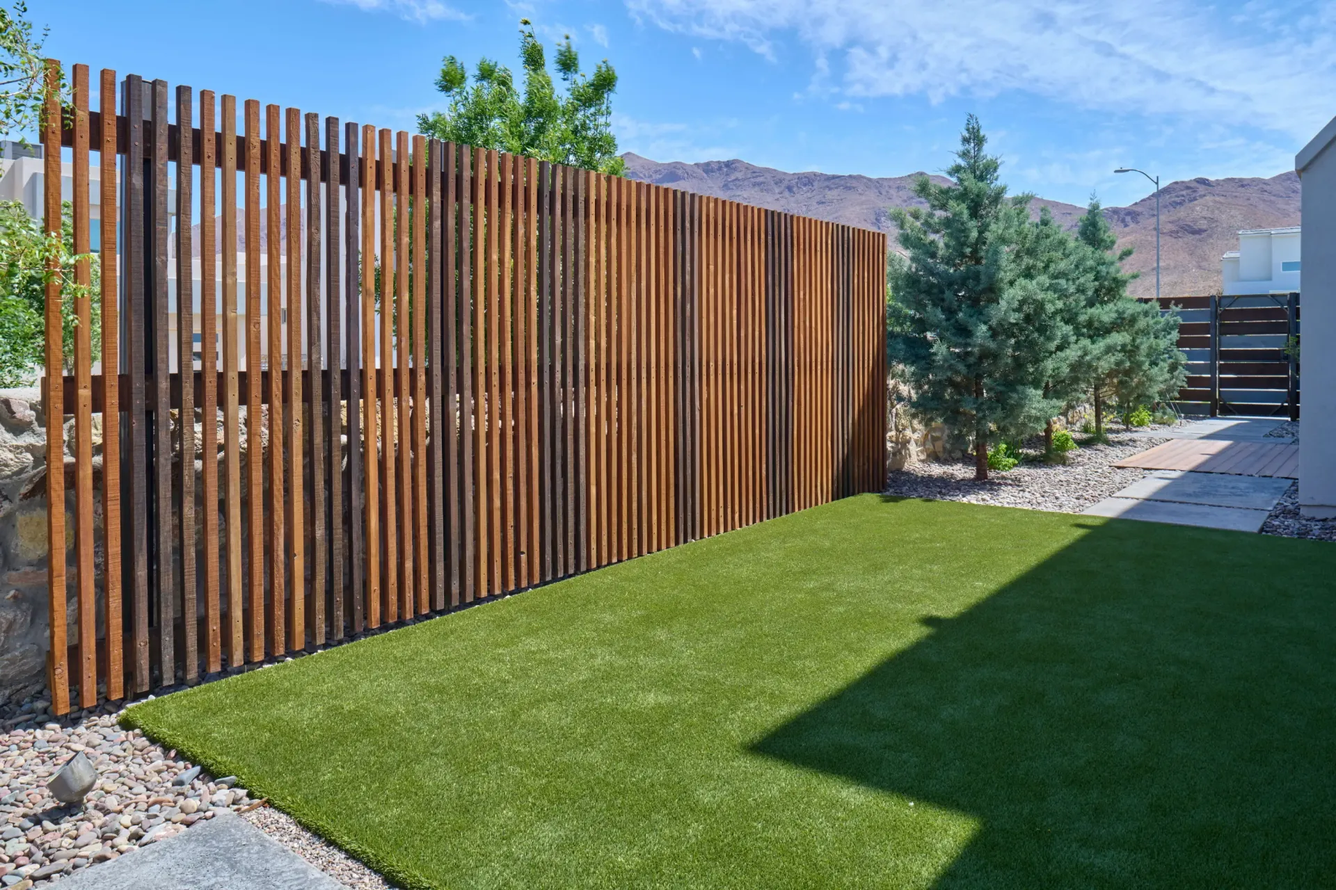 Artificial green lawn against a tall, wooden slatted fence. Trees and a walkway are visible.