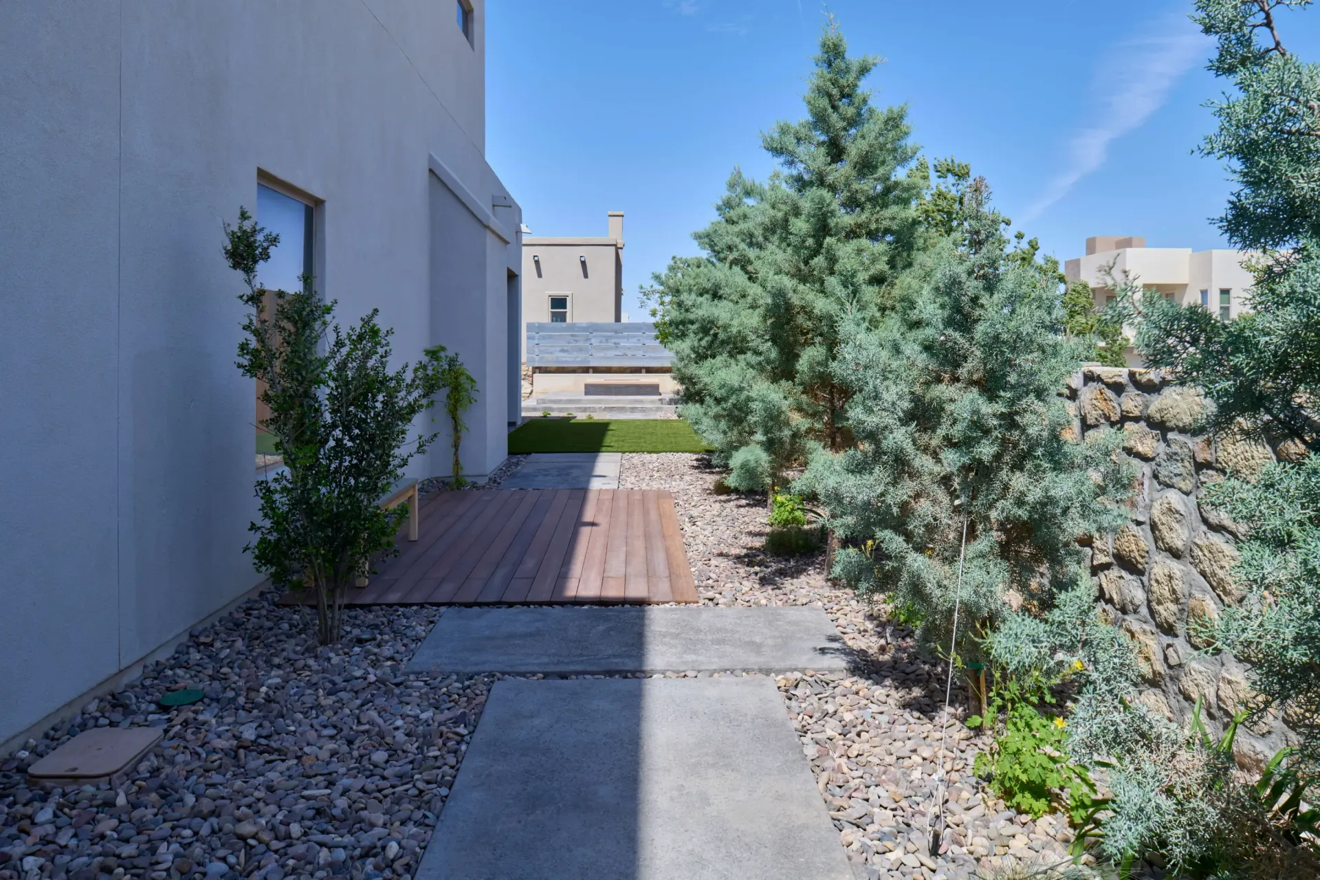 Path with wood and concrete sections, flanked by trees and gravel, leading toward building under a blue sky.