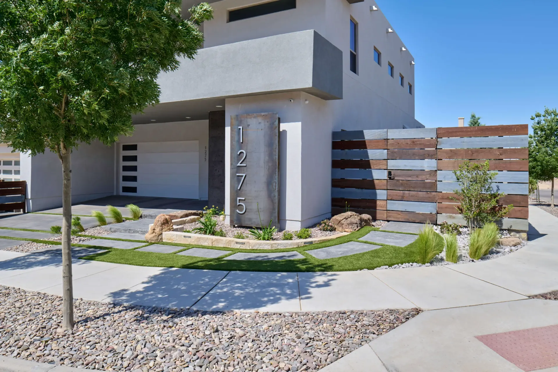 Modern two-story house with concrete accents, landscaping, and a wooden privacy fence on a sunny day. The address reads