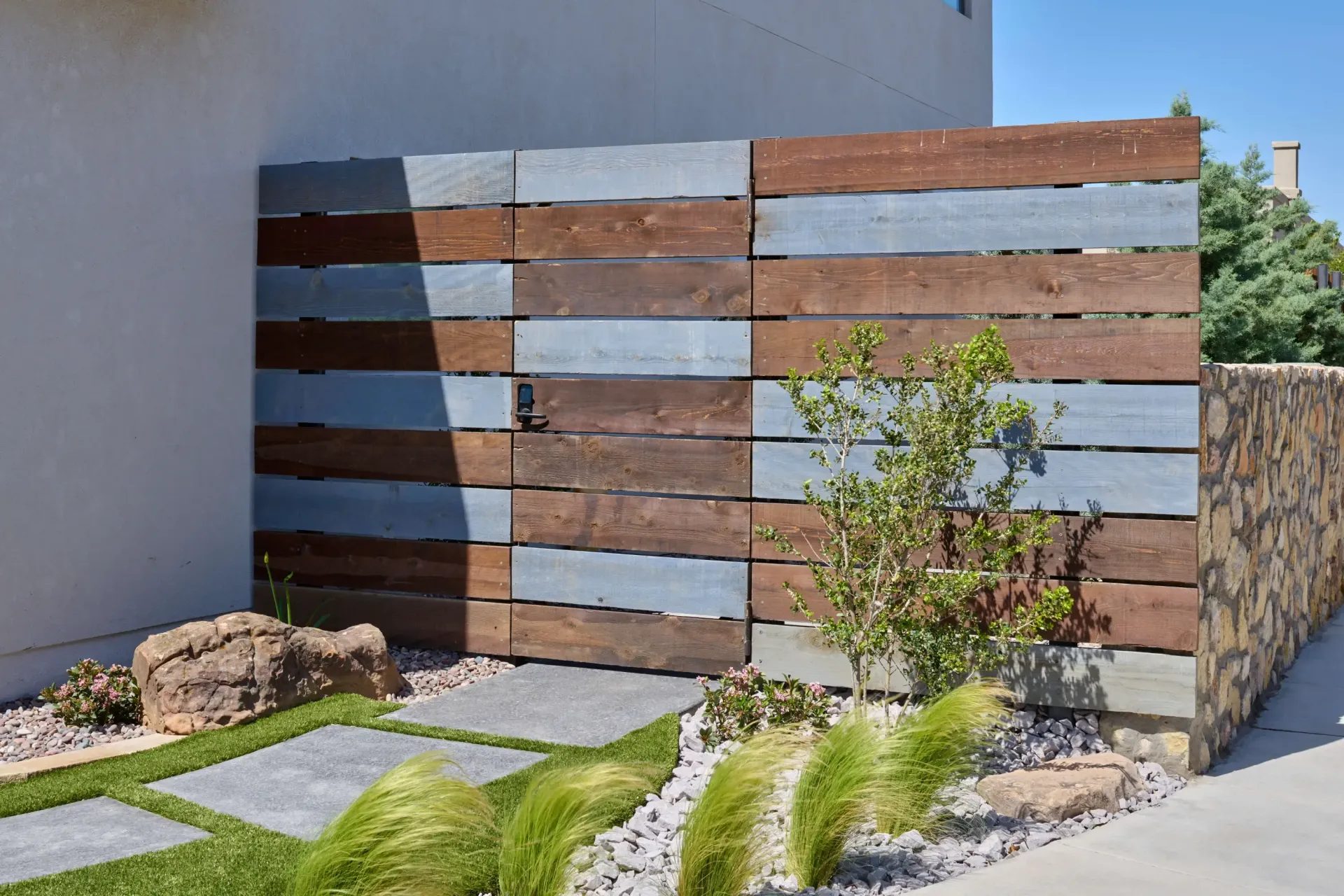 Wooden fence with brown and gray planks, next to a stone wall and small garden.