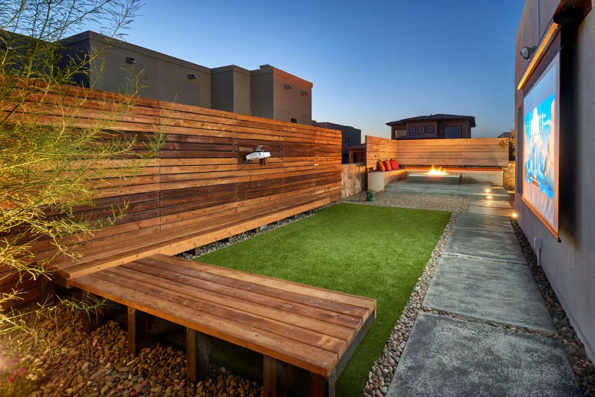 Outdoor entertainment area with wooden benches, faux grass, fire pit, and a screen; evening.