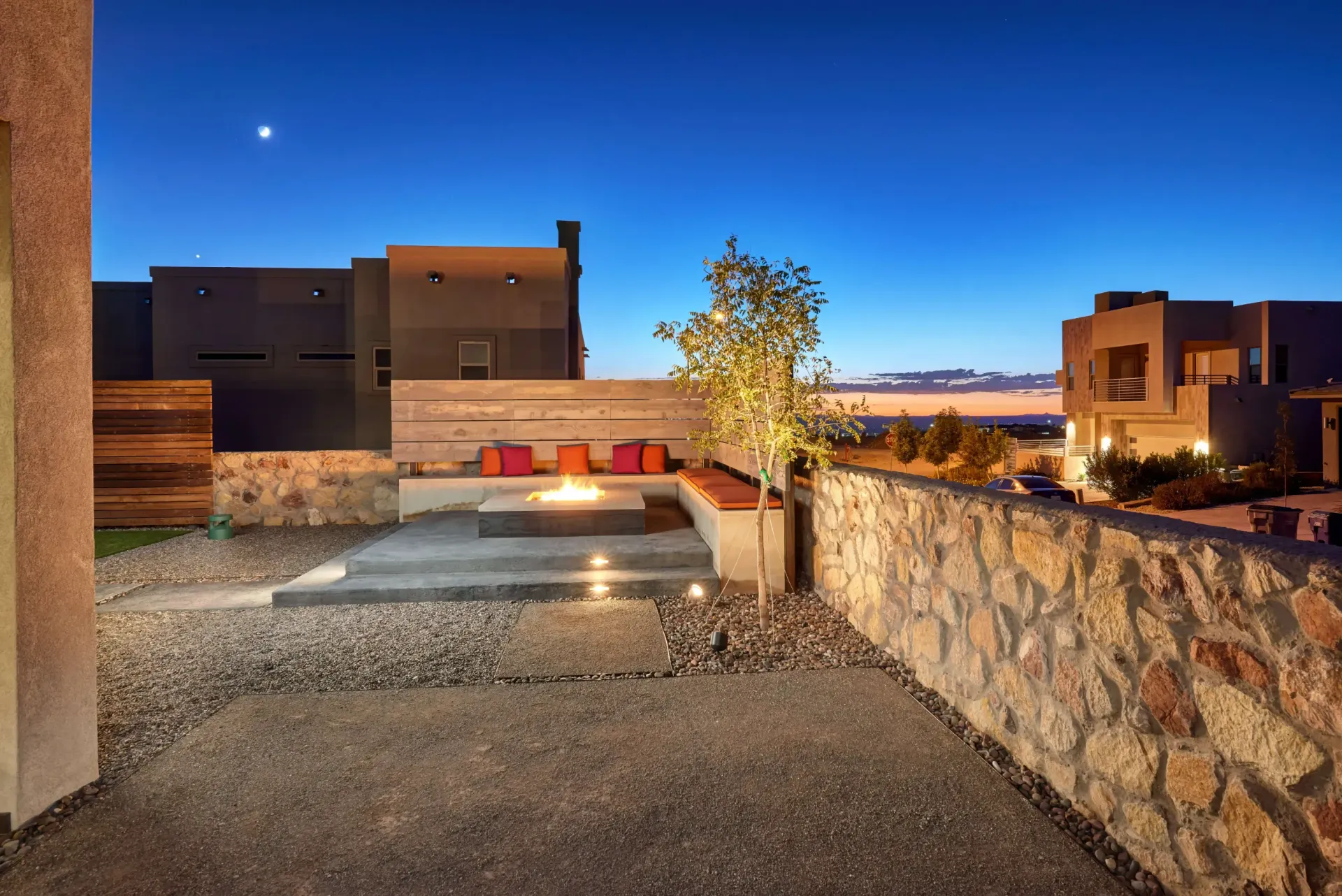 Outdoor patio at dusk with fire pit, seating, stone wall, gravel, and homes in the background.