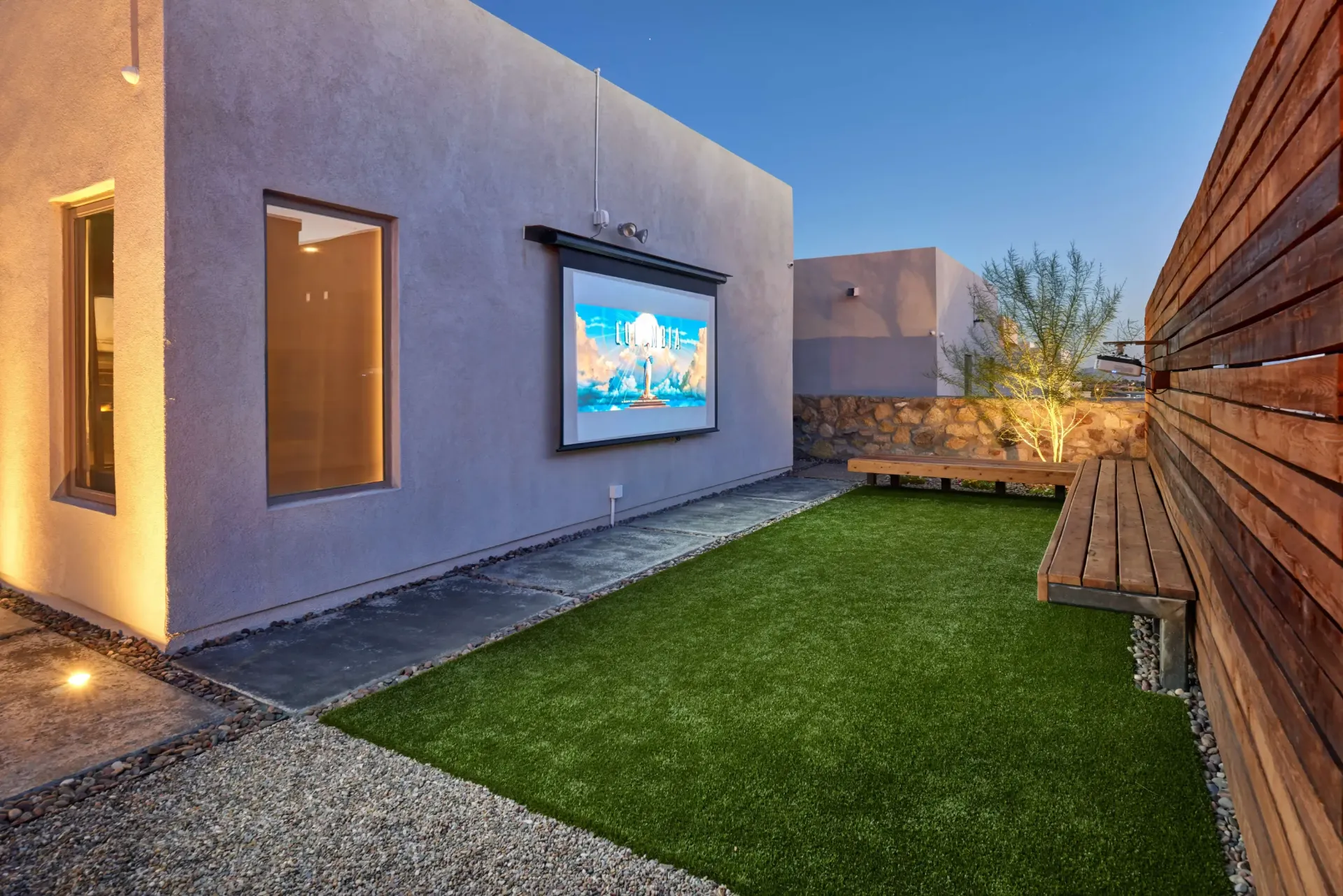 Outdoor entertainment area with a projector screen on a building, grass, wooden benches, and a gravel pathway.