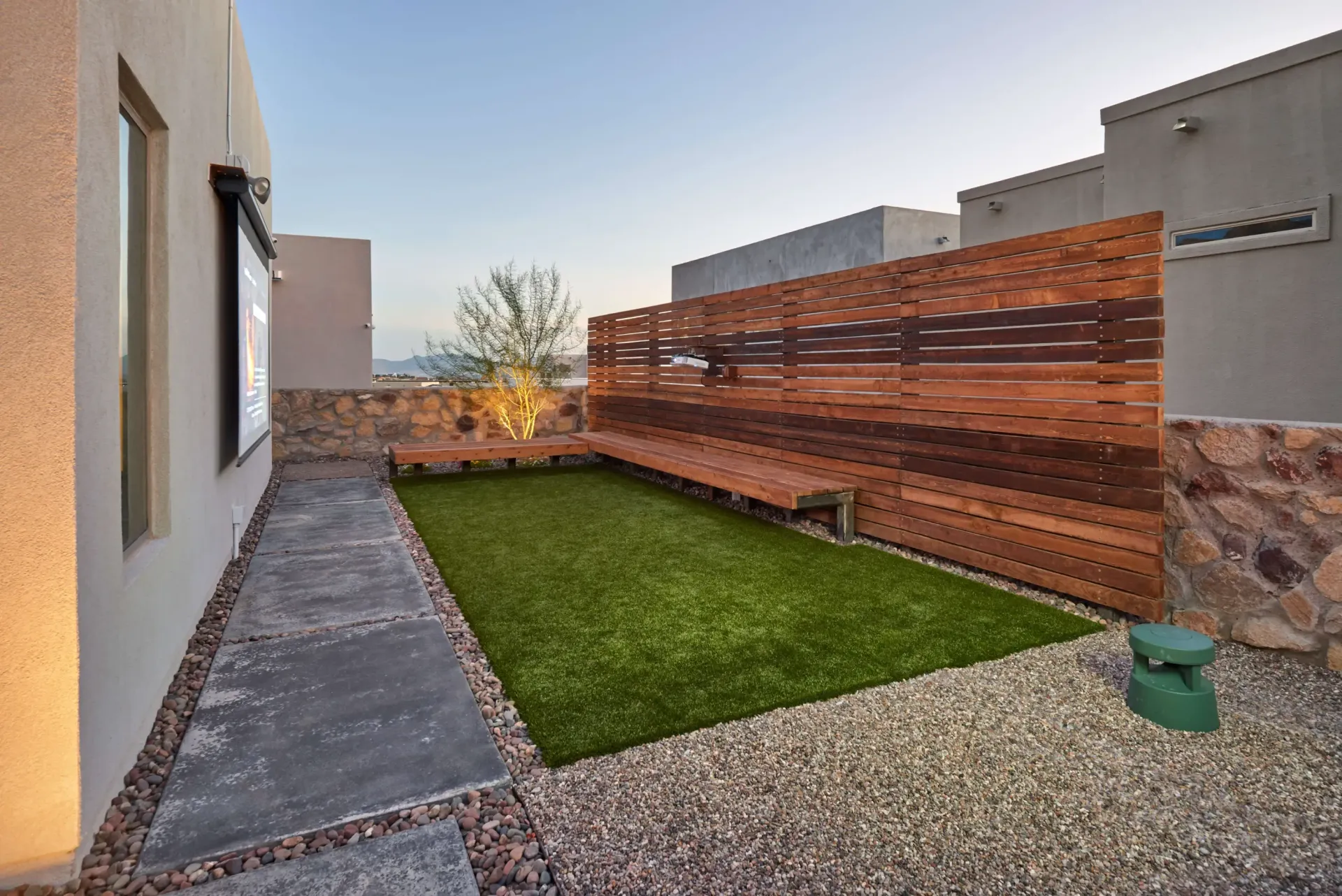 Rooftop patio with artificial grass, wooden screen, and seating, surrounded by gravel and beige buildings.