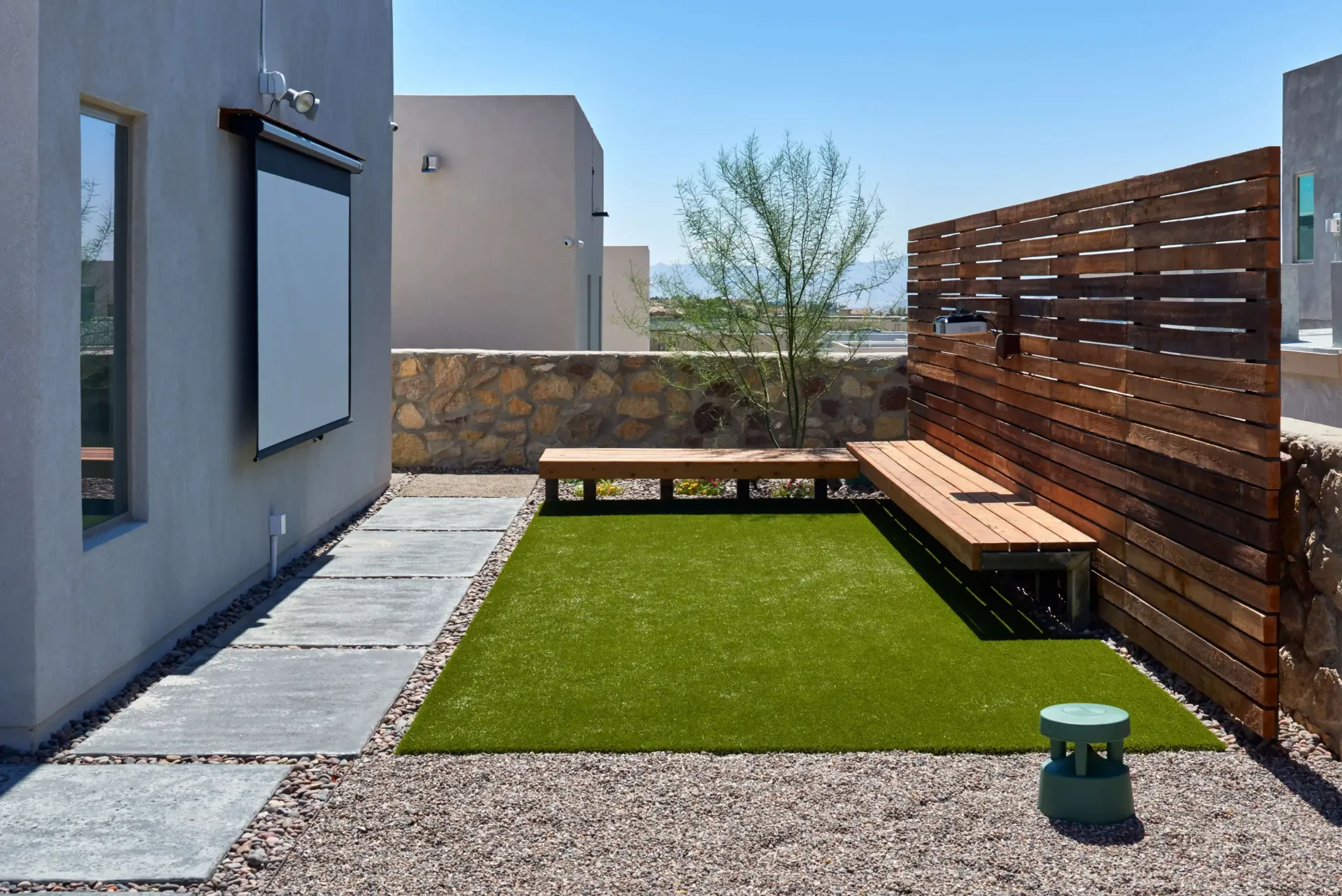 Small backyard with green turf, wooden benches, and a screen; sunny day.