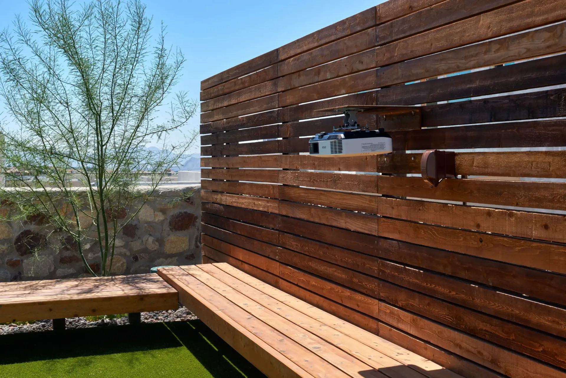 Wooden bench and slatted fence in outdoor space, with tree and rock wall in the background.