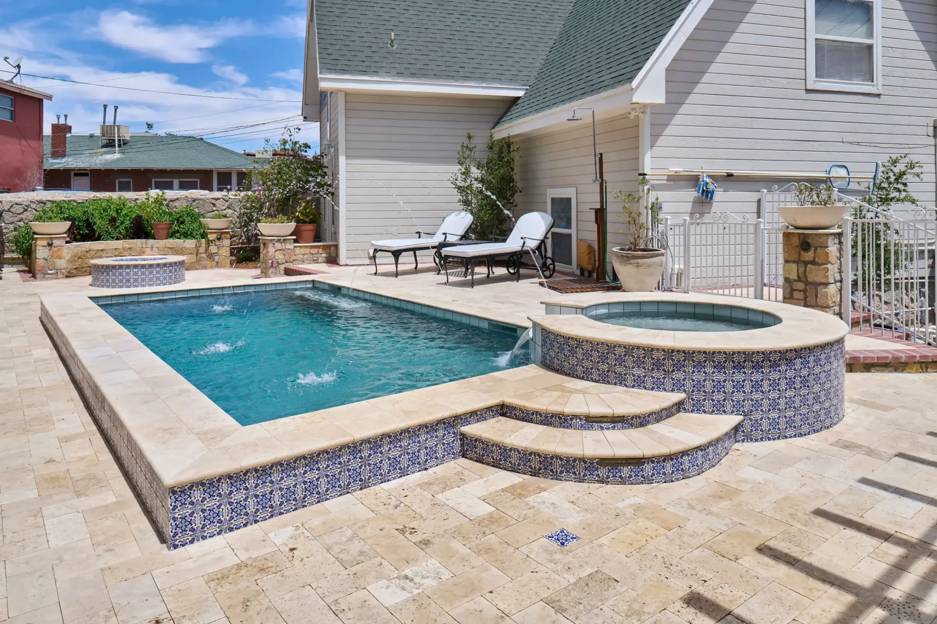 Backyard with rectangular pool and circular hot tub, beige paving, and a white house.
