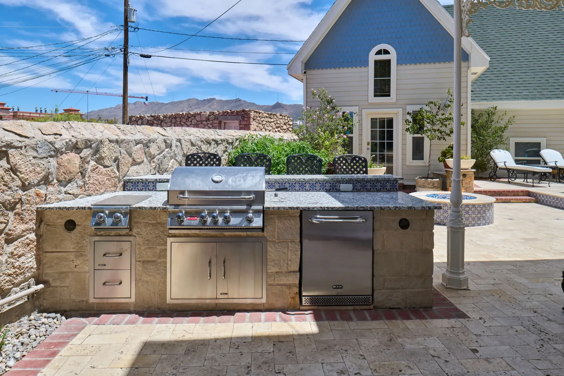 Outdoor kitchen with grill, cooktop, and dishwasher set against a blue house and stone wall.