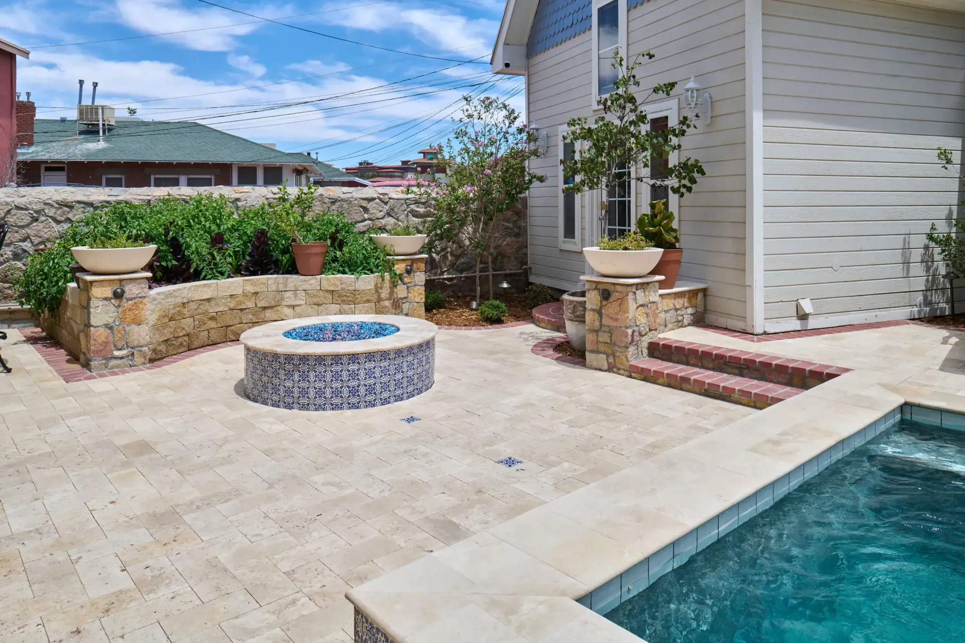 Patio with fire pit, stone walls, pool, and house under a bright blue sky.