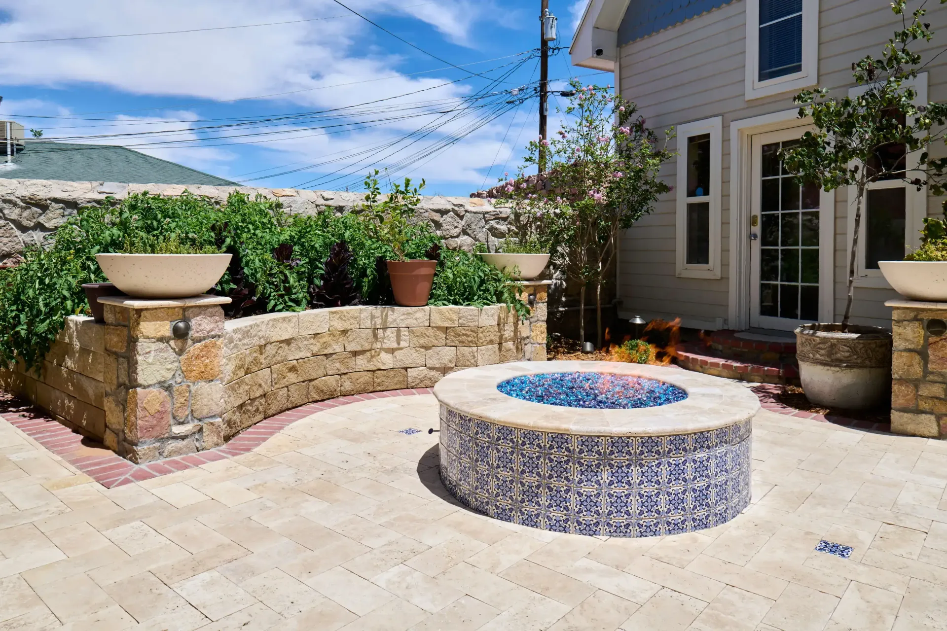 Patio with mosaic fire pit, stone wall, potted plants, and light blue house against a partly cloudy sky.