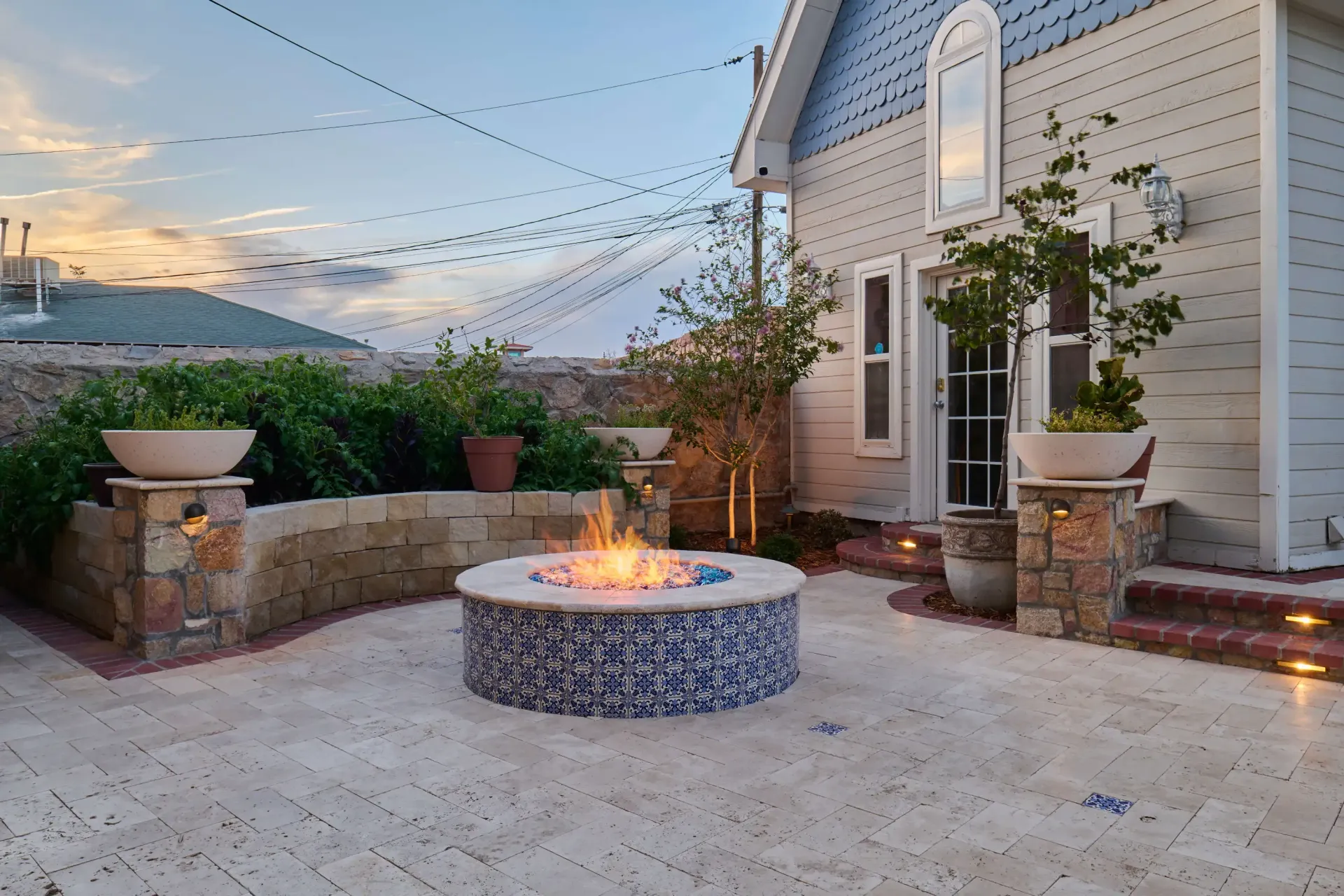 Outdoor patio with fire pit, stone wall, potted plants, and string lights, near a light blue building.