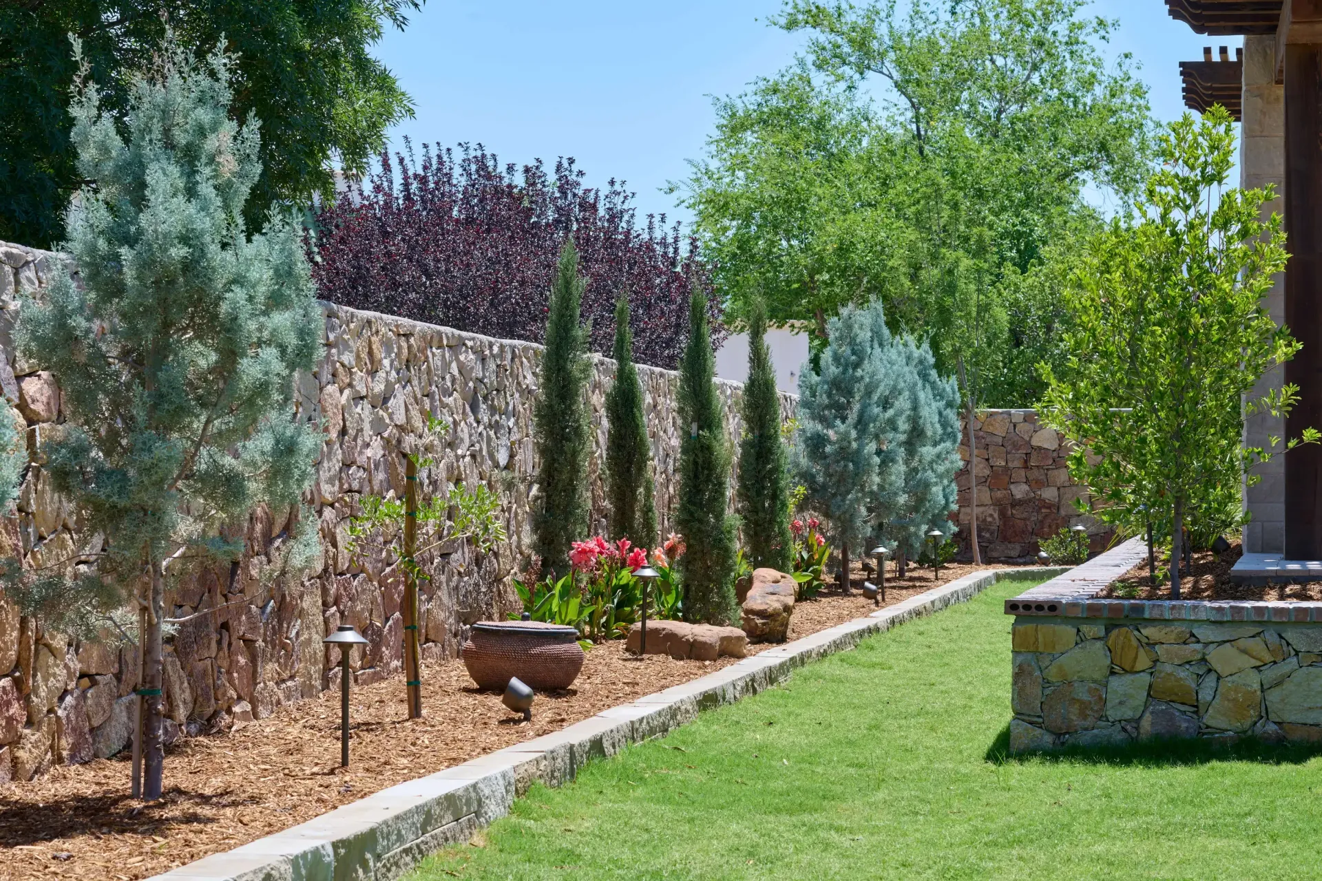 Stone wall borders a backyard with cypress and blue spruce trees, green grass.