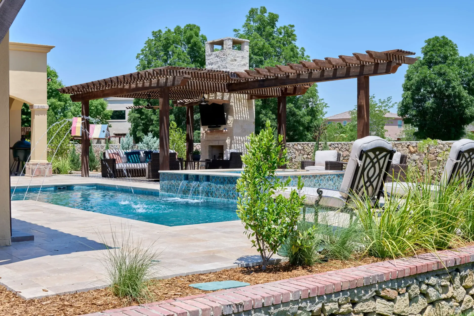 Swimming pool with a pergola and outdoor fireplace. Lush greenery, blue sky, and stone accents.