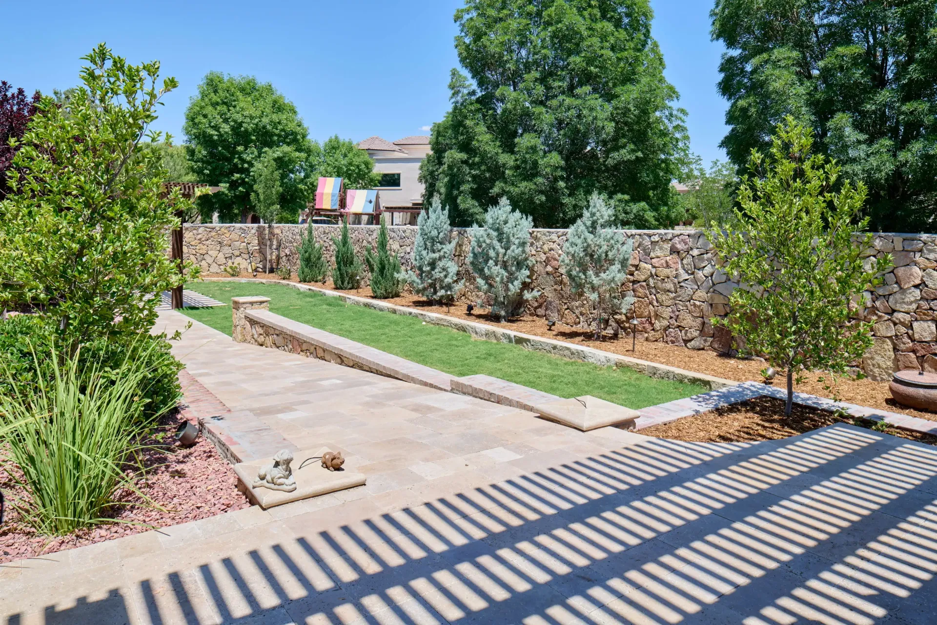 Stone-walled backyard with green grass, small trees, and concrete paths; sunny day with shade from overhead structure.