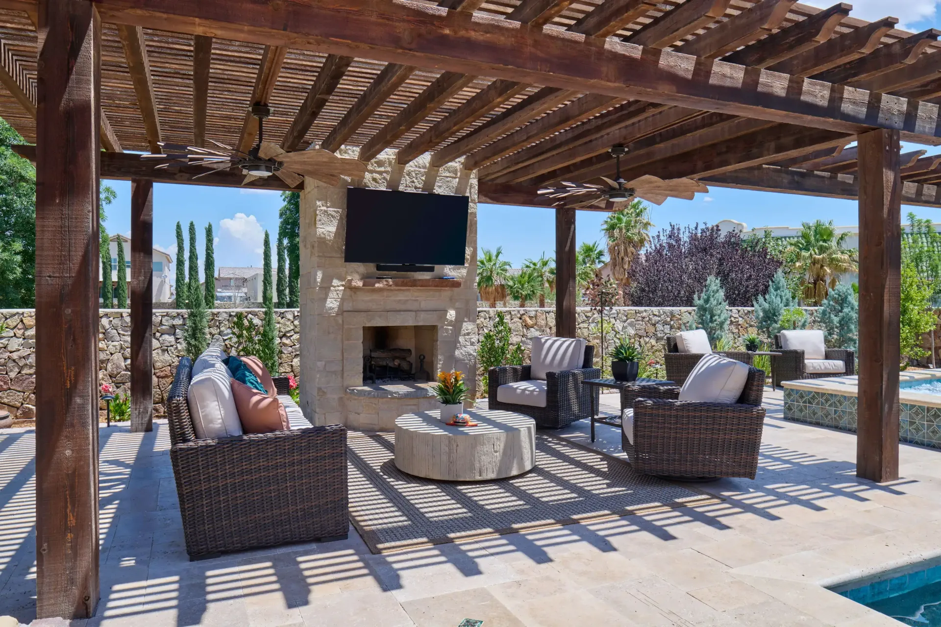 Outdoor living space under a wooden pergola with fireplace, TV, and seating, overlooking a pool and landscaping.
